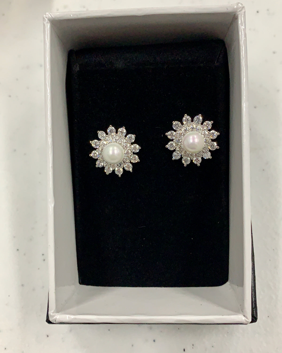 A pair of silver-toned flower-shaped stud earrings with white pearl centers and clear stones, presented in a white jewelry box with a black velvet insert.