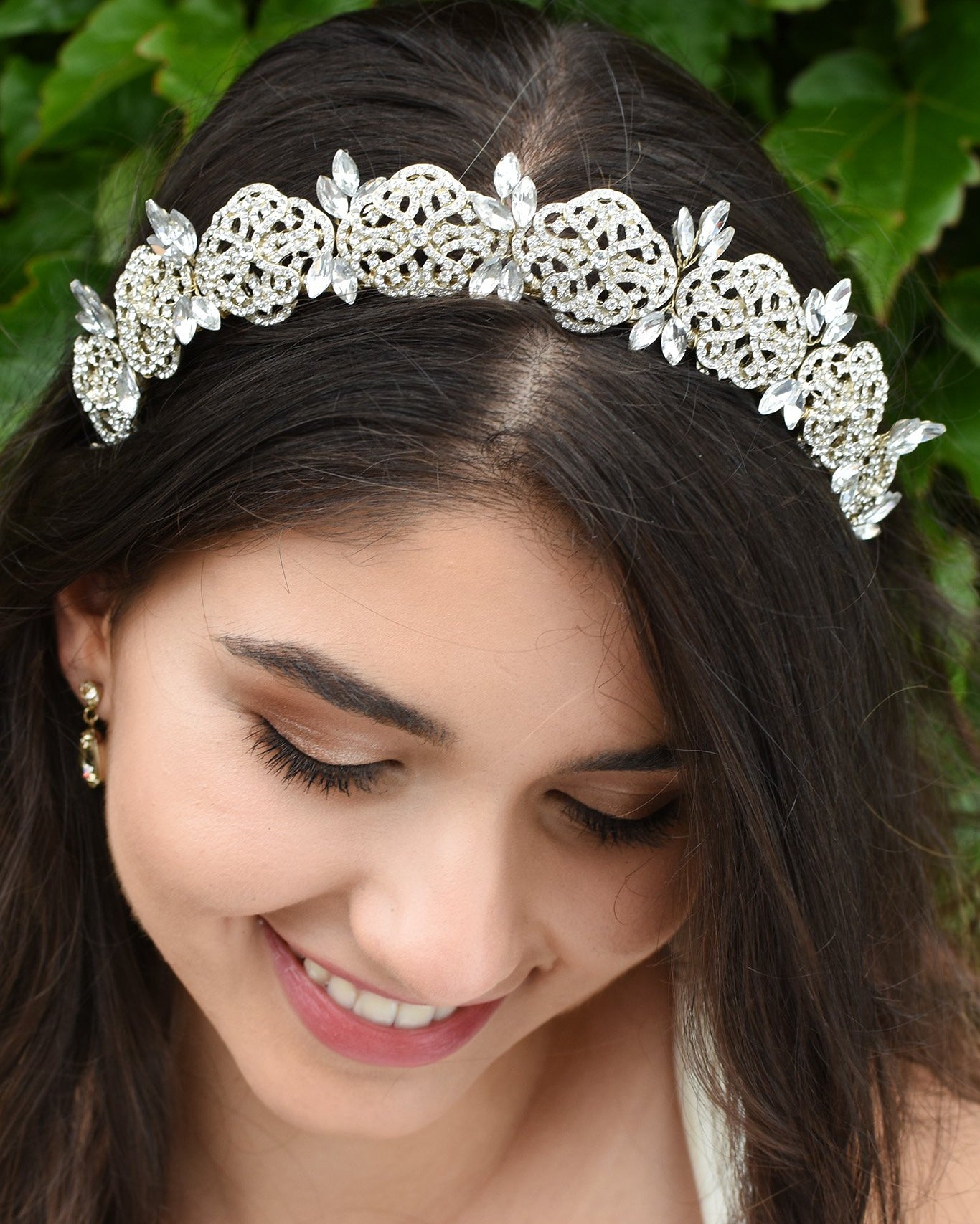 Smiling Bride wears a low tiara with crystals in pale gold. She has dark hair and green leaves behind her.
