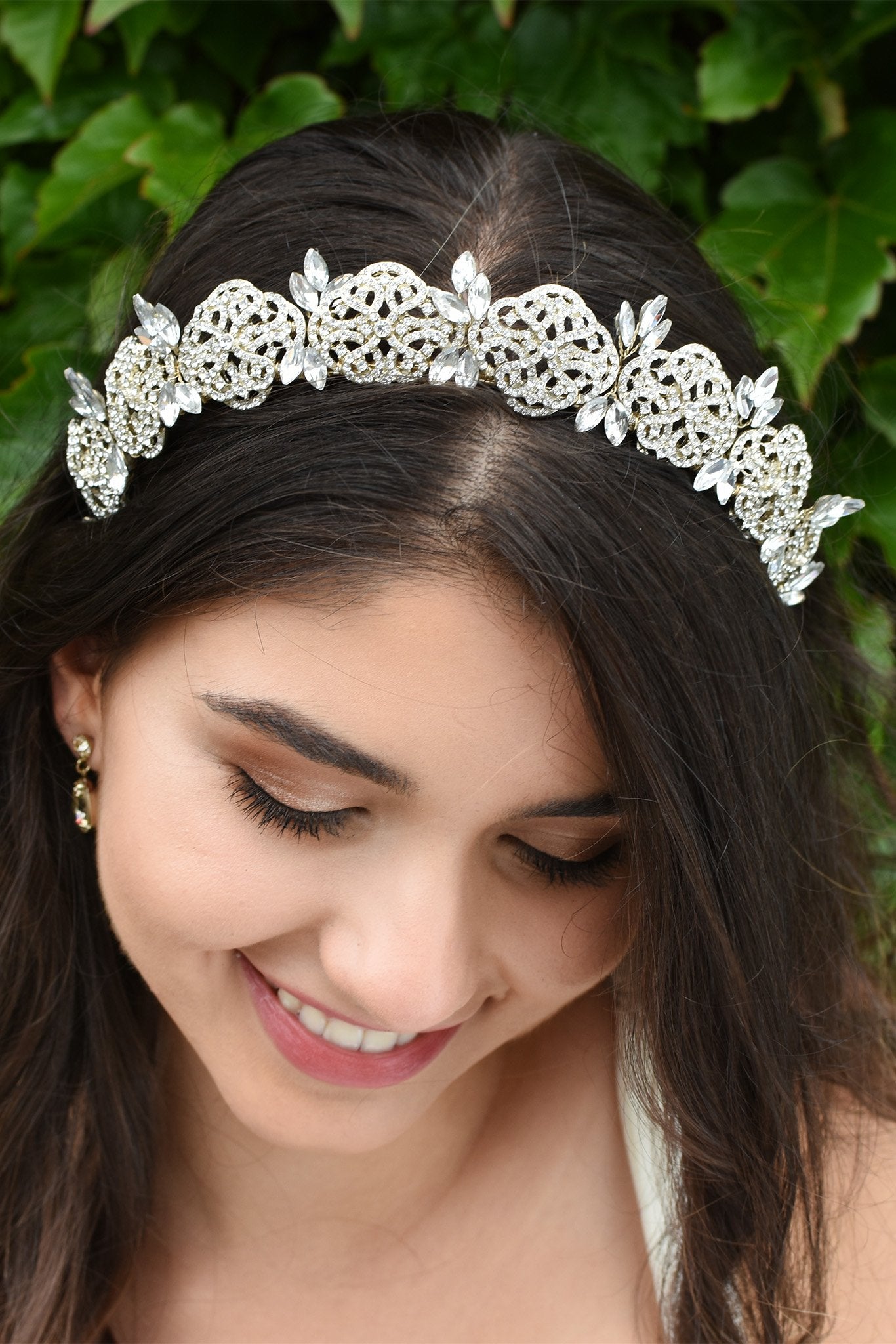Smiling Bride wears a low tiara with crystals in pale gold. She has dark hair and green leaves behind her.