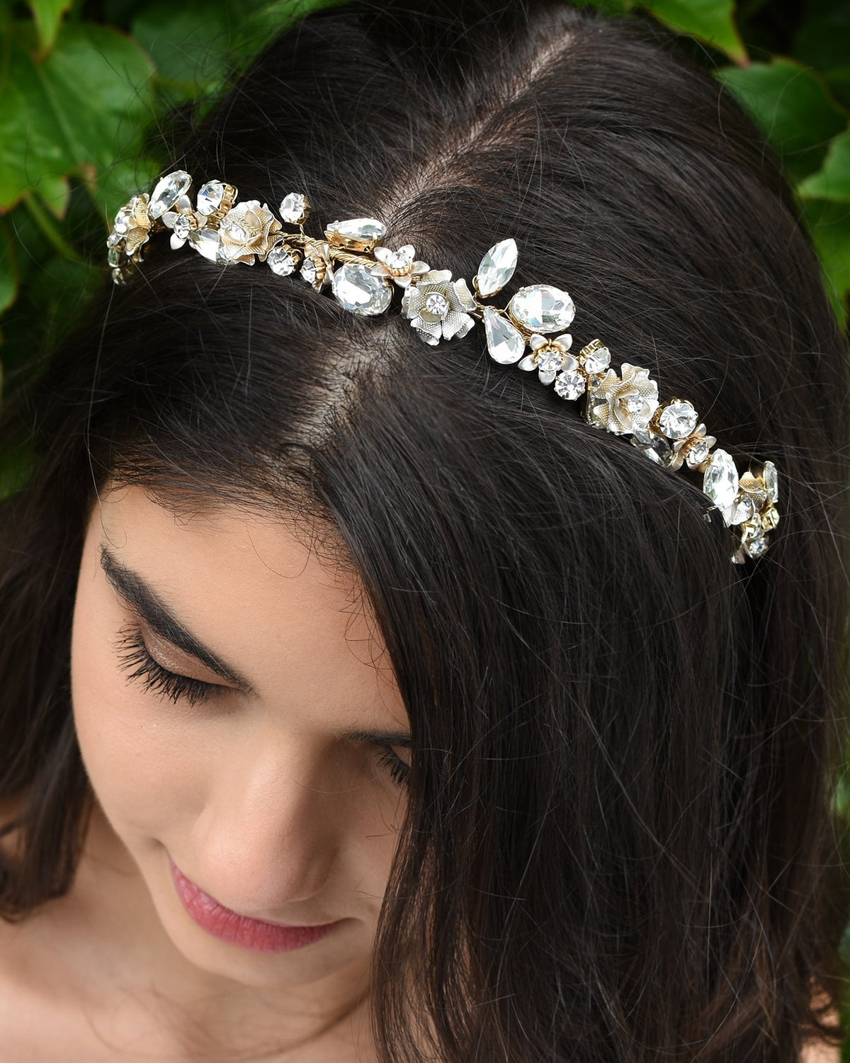Dark hair smiling Bride wearing a thin headband full of different shapes of stones with green leaves in the background