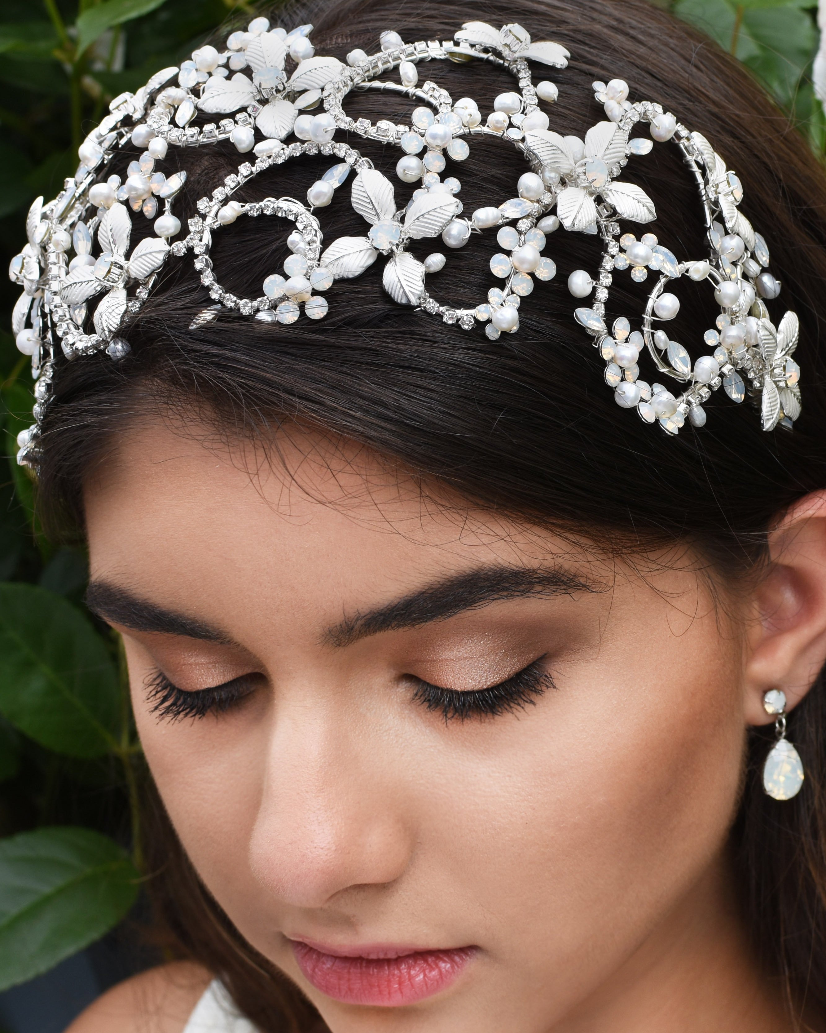 Dark haired model wears a very wide silver headband on the front of her head with green leaves as a background