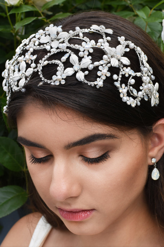 Dark haired model wears a very wide silver headband on the front of her head with green leaves as a background