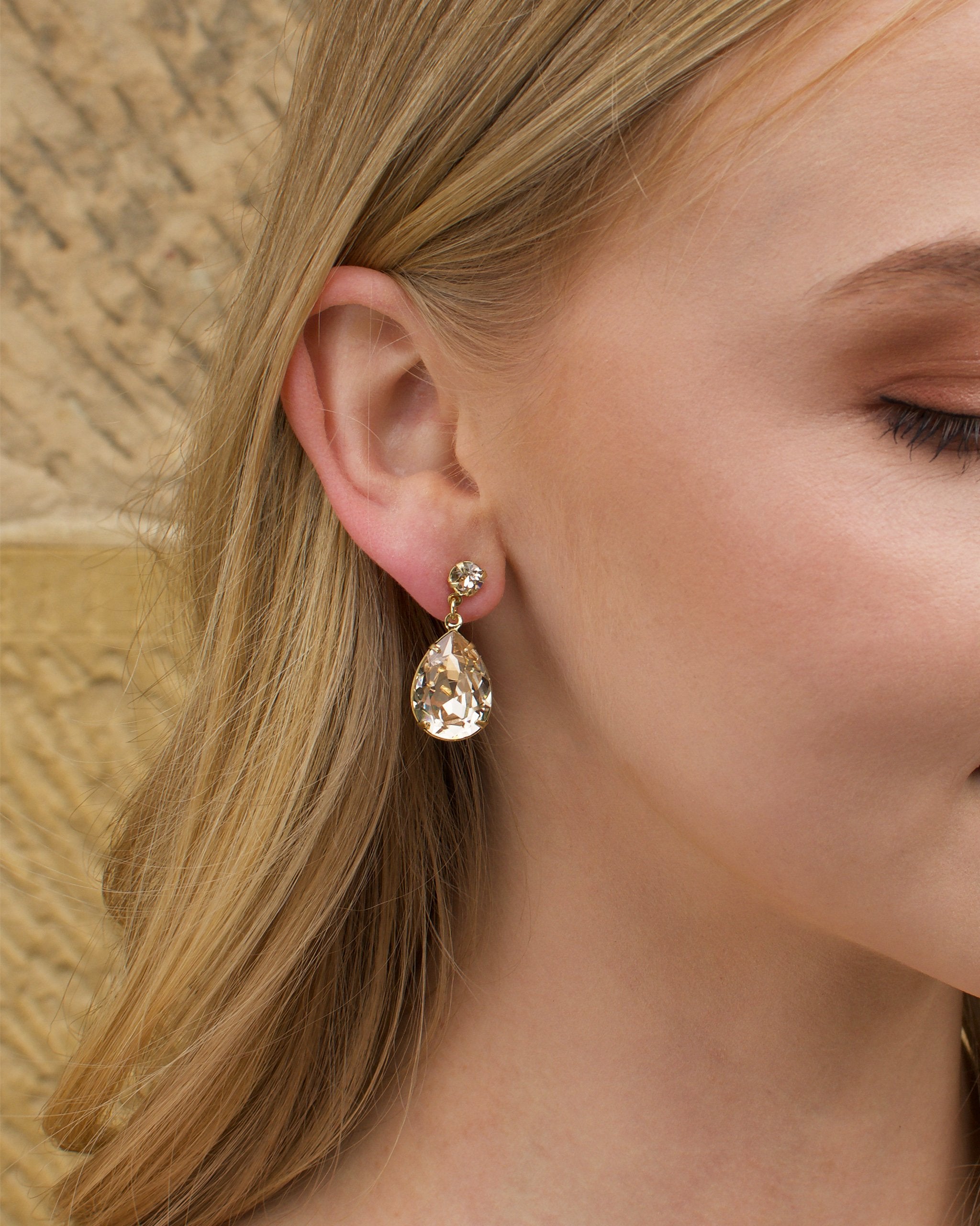 A blonde model wears a pale gold earring with a silk colour stone in her ear with a stone wall background