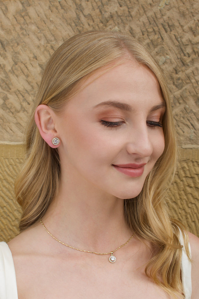 A blonde hair model wears a gold stud earring with a simple gold chain with a stone wall backdrop