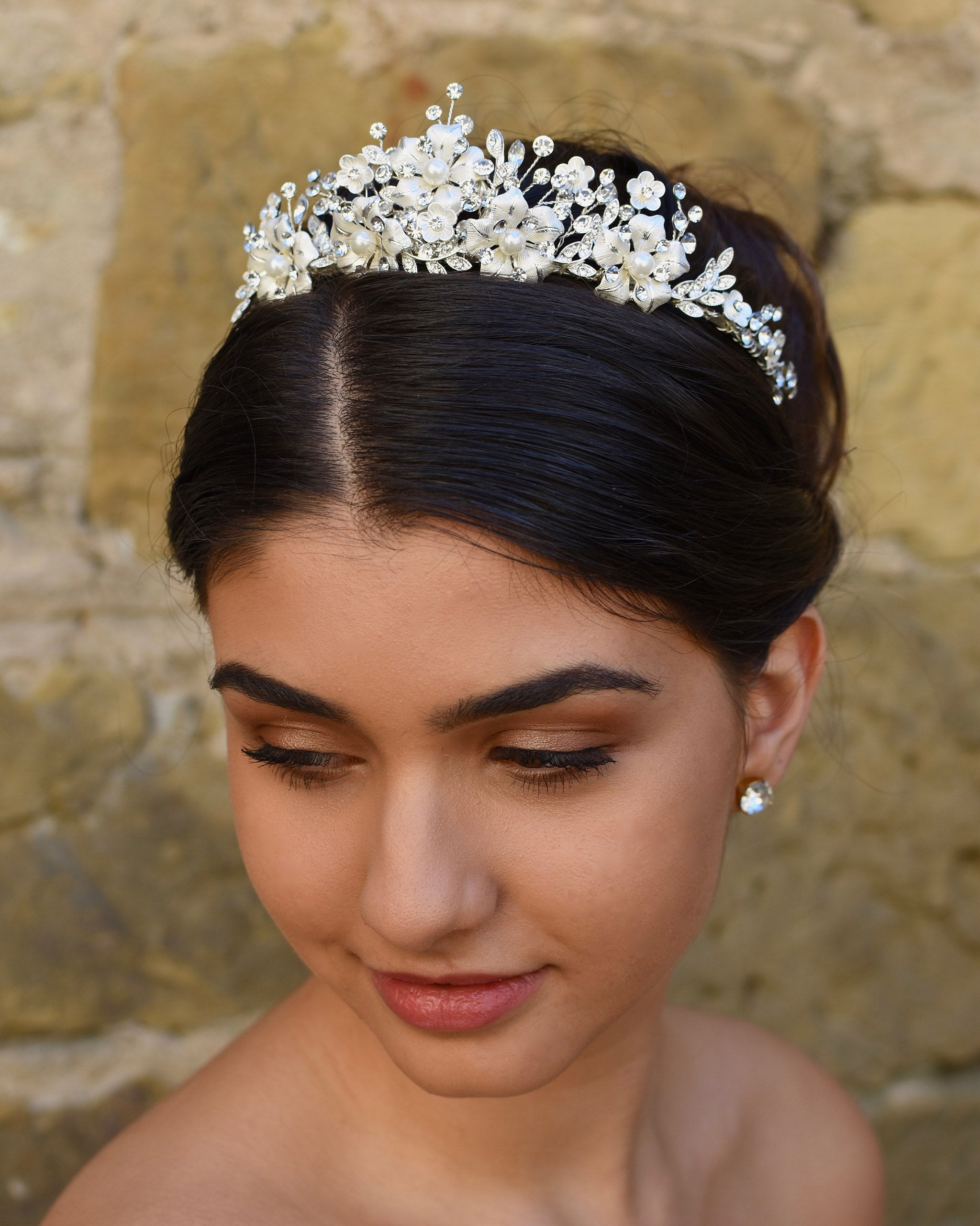 A dark haired model wears a silver tiara with pearl flowers against a stone wall