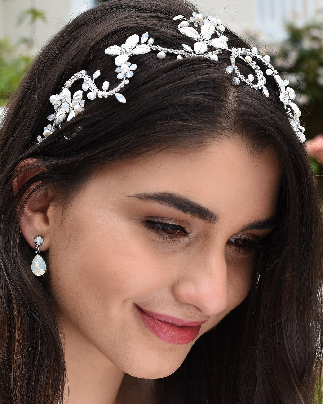Dark Haired smiling model wears a silver bridal headband with white opal stones with a garden background