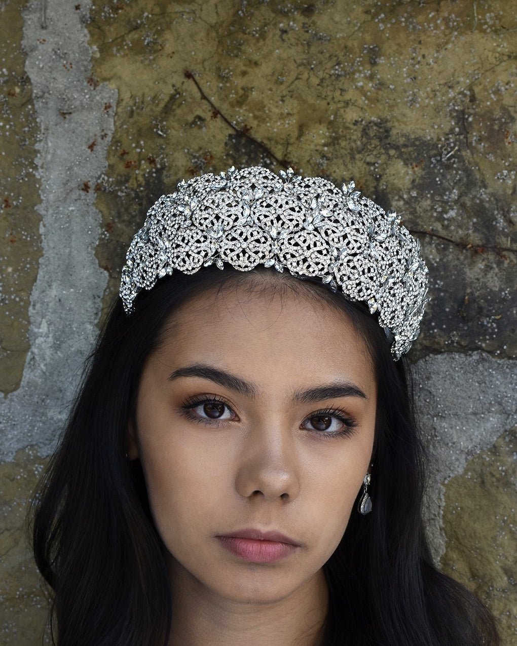The front view of a bride wearing a high Bridal Tiara covered with tiny crystals with a stone wall background