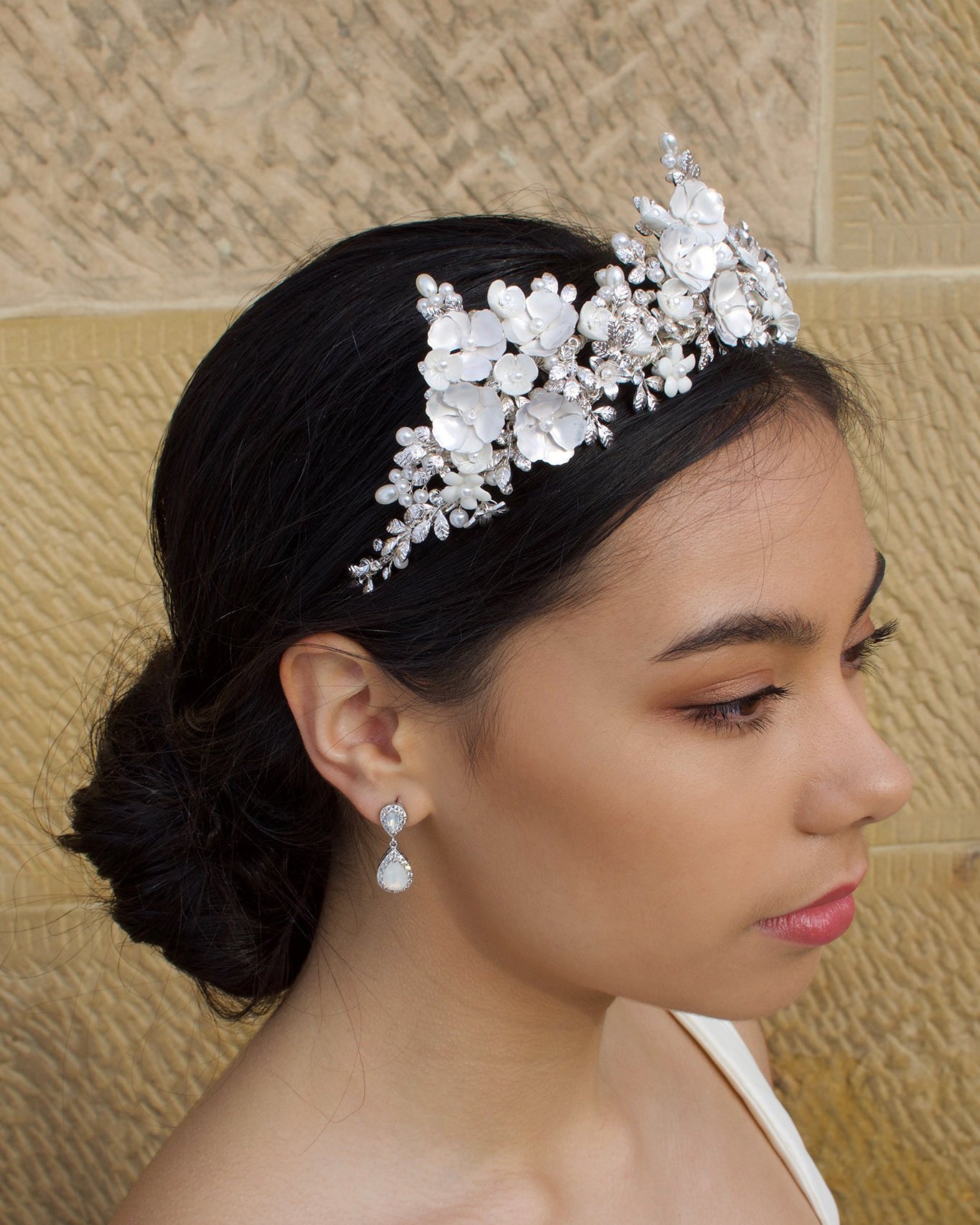 A dark haired bride wears a three pointed flower tiara at the front of her head. Behind is a sandstone wall.