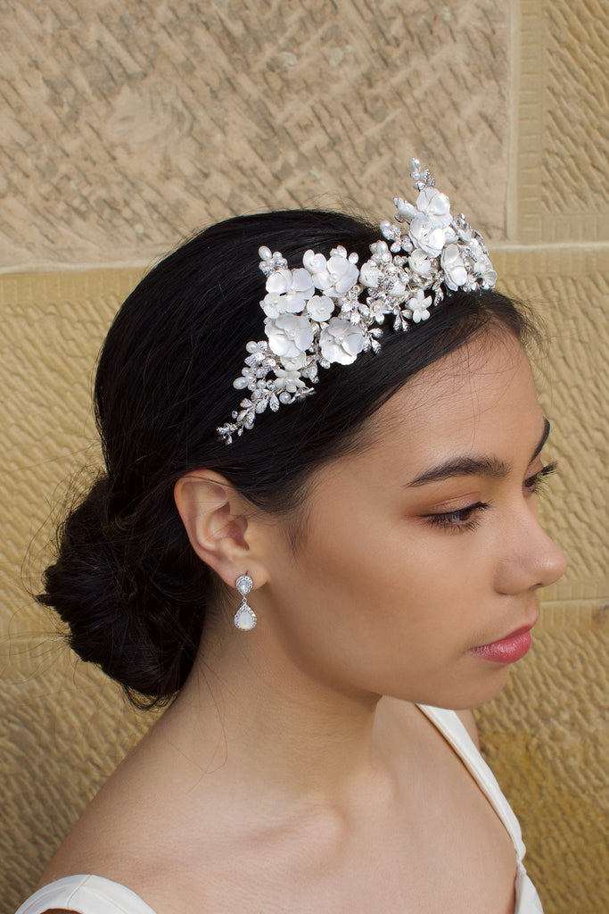 A dark haired bride wears a three pointed flower tiara at the front of her head. Behind is a sandstone wall.