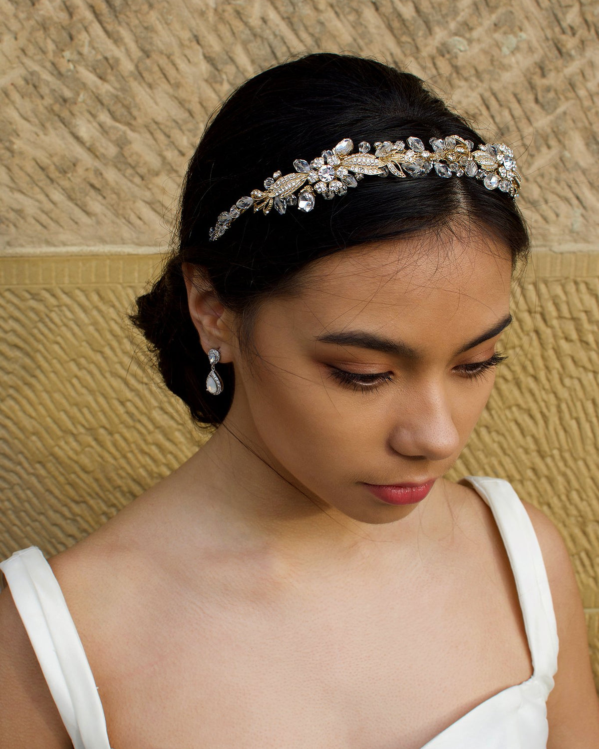 Dark haired model with long hair wears a side comb of pale gold parts at the front of her head. With a stone wall background