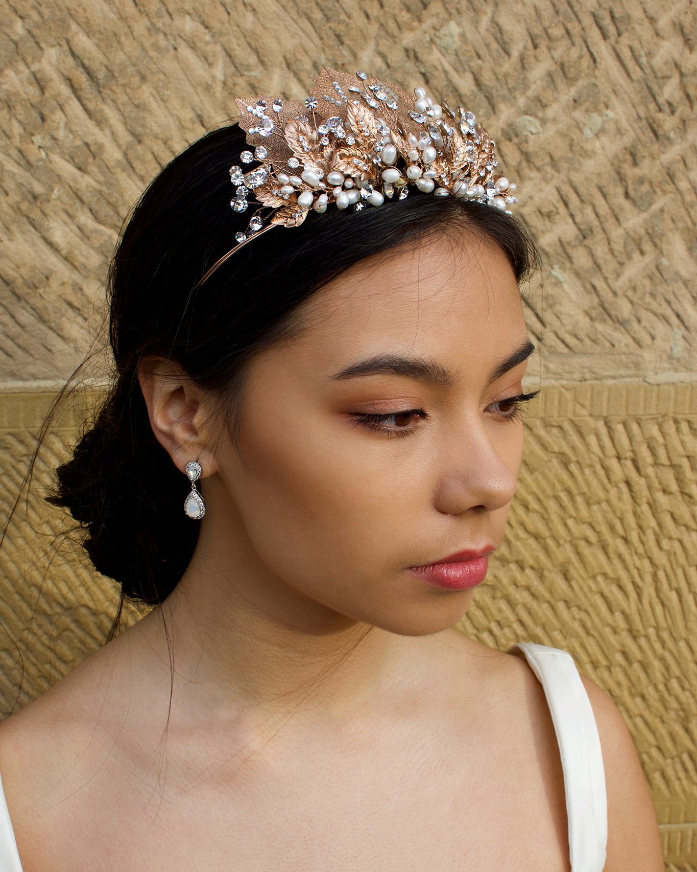 A black haired model wears a rose gold tiara with pearls in the background is a wall.
