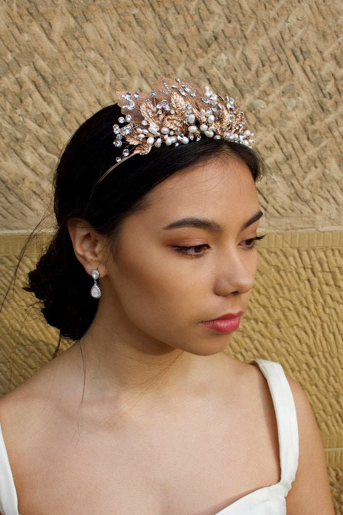 A black haired model wears a rose gold tiara with pearls in the background is a  wall.