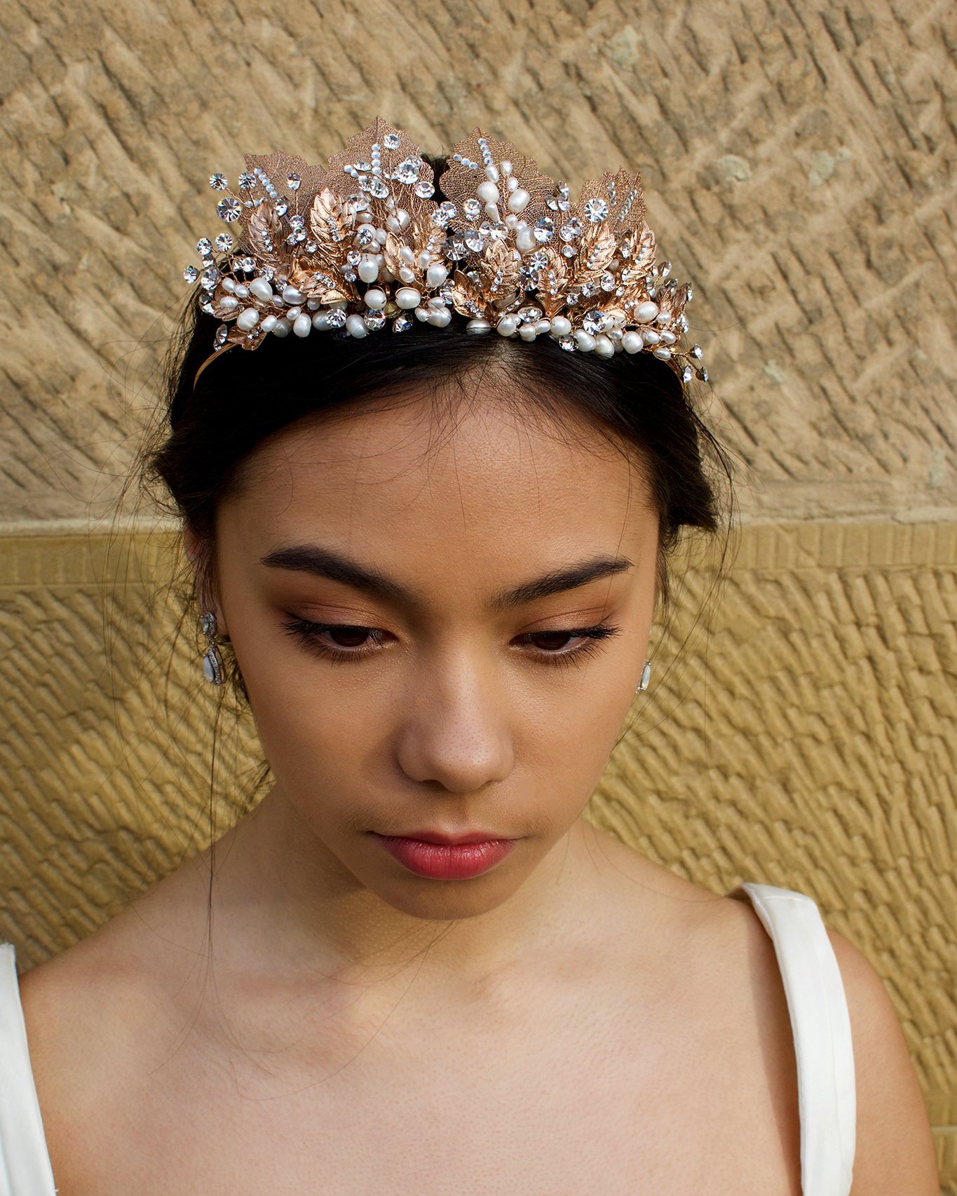 A black haired model wears a rose gold tiara with pearls in the background is a sandstone wall.