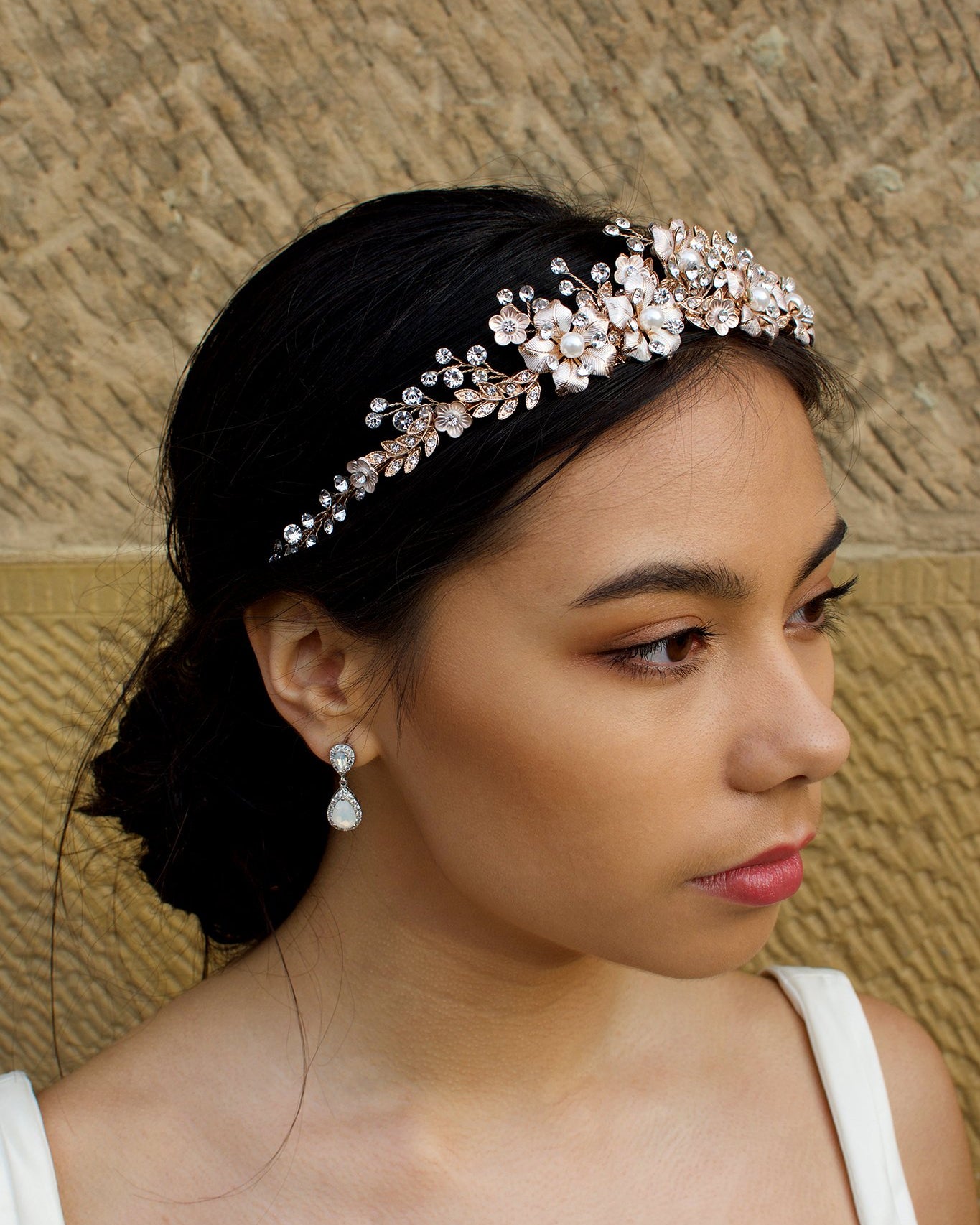 A dark haired model wears a rose gold tiara with pearl flowers against a stone wall background.
