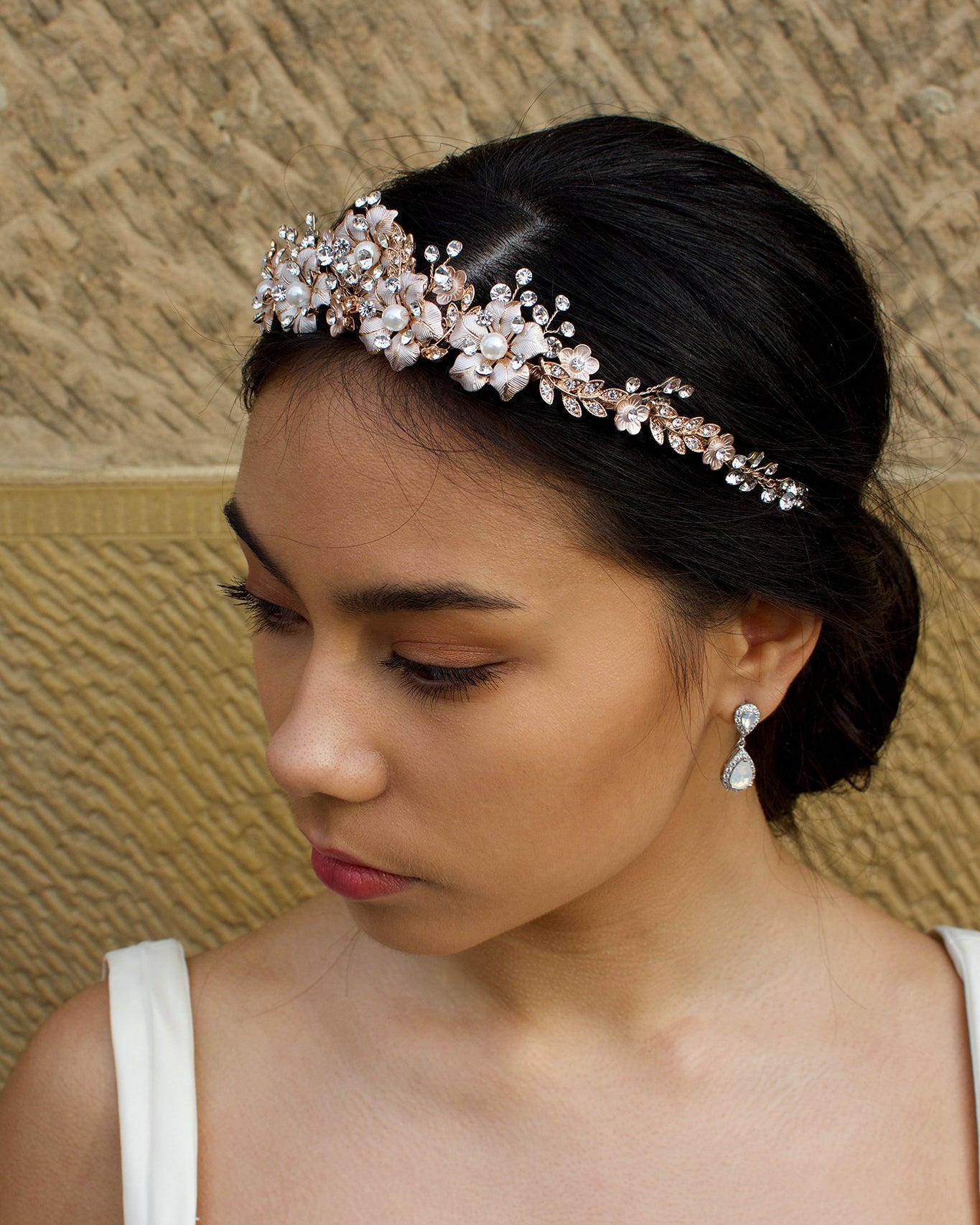 A dark haired model wears a rose gold tiara with pearl flowers against a stone wall background.
