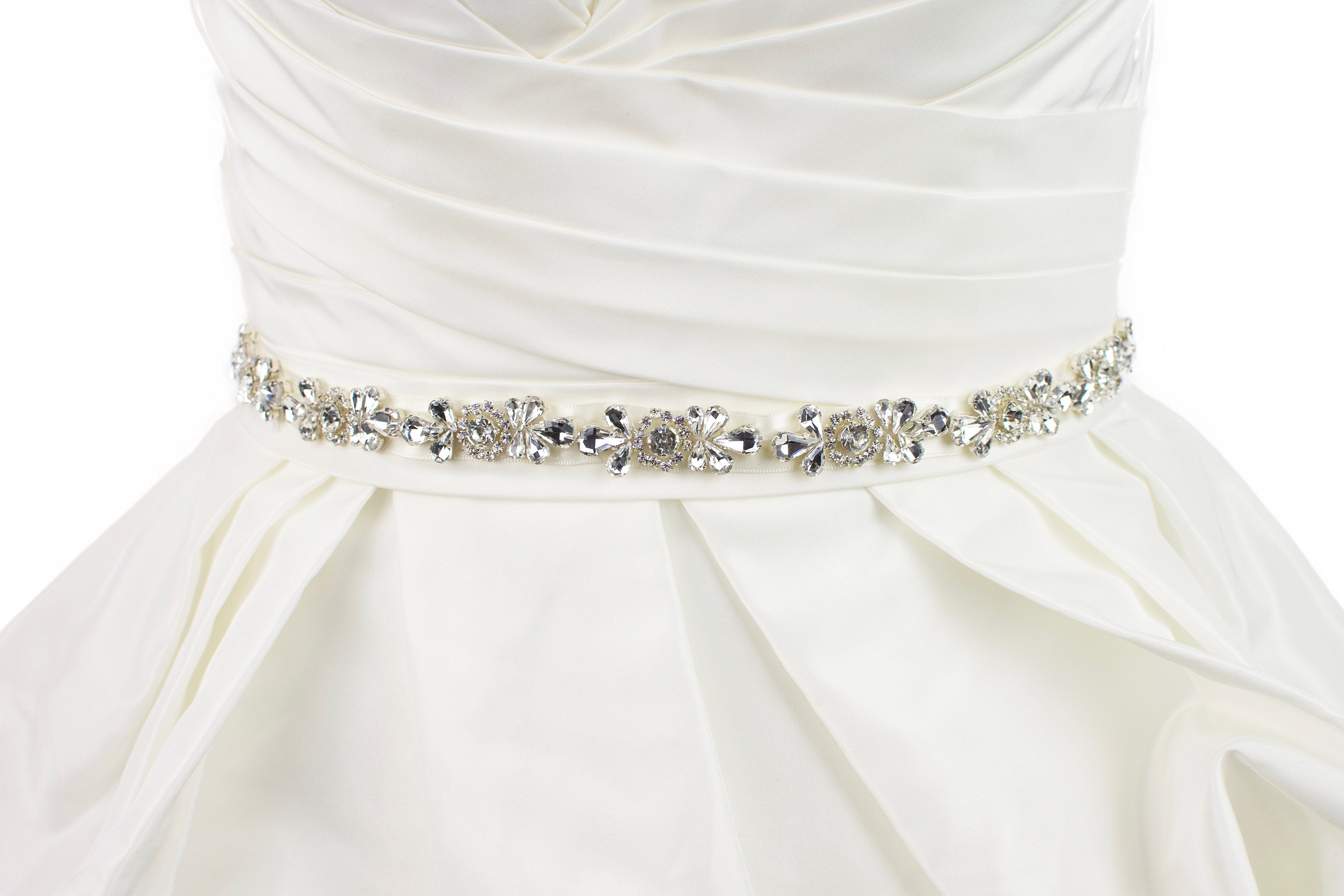 A belt of teardrop shape crystals is around the waist of an ivory satin wedding gown with a white background.