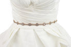 A narrow rose gold belt with crystals is fitted around the waist of an ivory bridal gown with a white background
