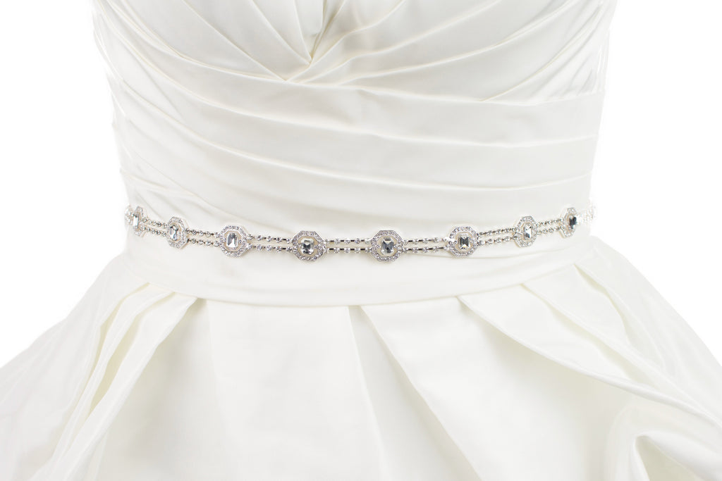 A narrow silver chain bridal belt is fitted around the waist of an ivory bridal gown with a white background