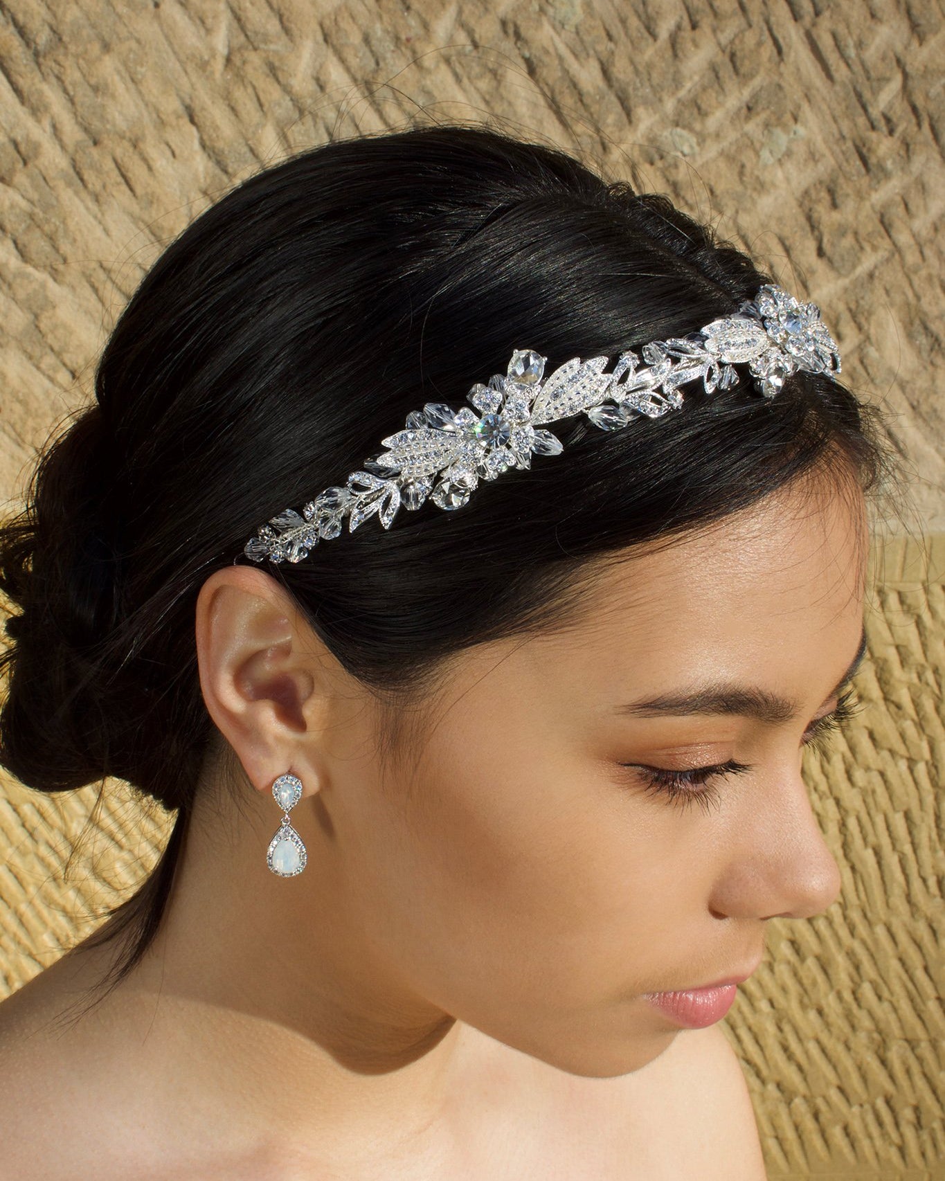 A dark haired model wears a Swarovski crystal crystal headband on the front of her head. The background is a stone wall.