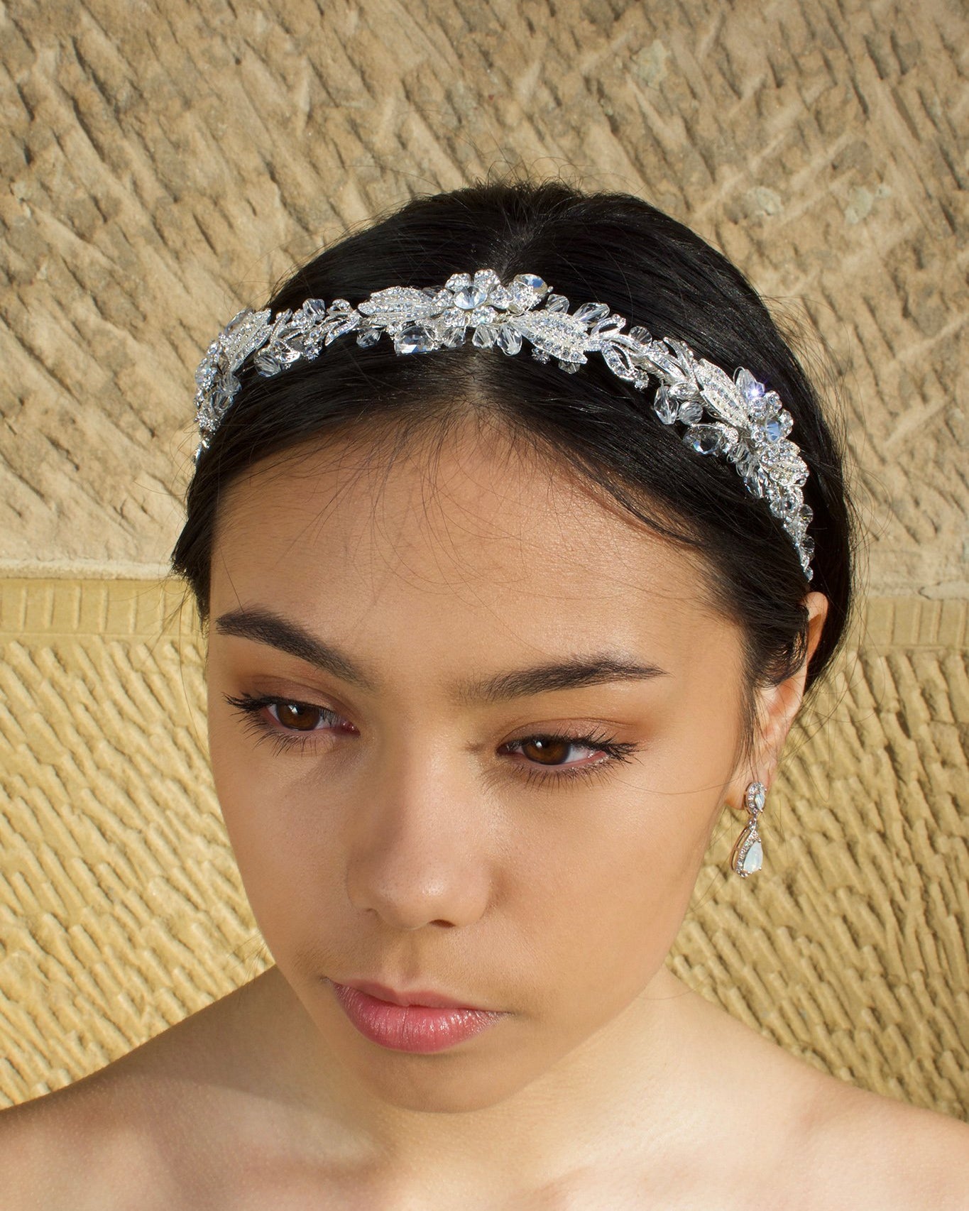 Black haired model with long hair wears a side comb of pale gold leaves at the side of her head. With a stone wall background
