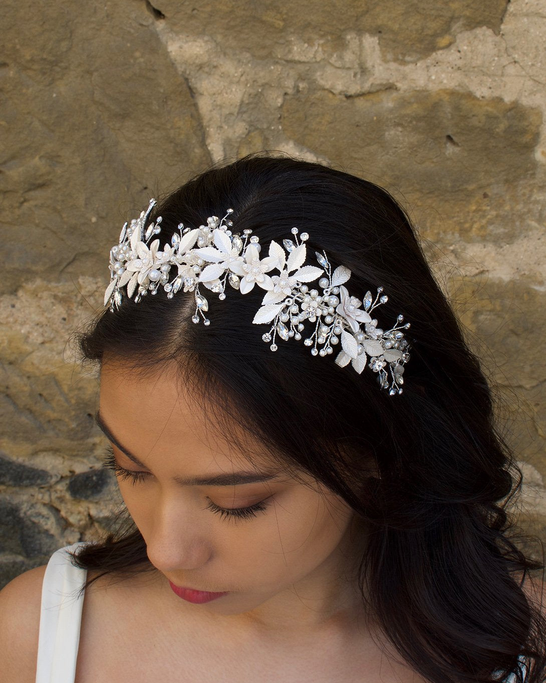 Model looks down wearing a silver leaves headband on her dark hair with an old wall behind