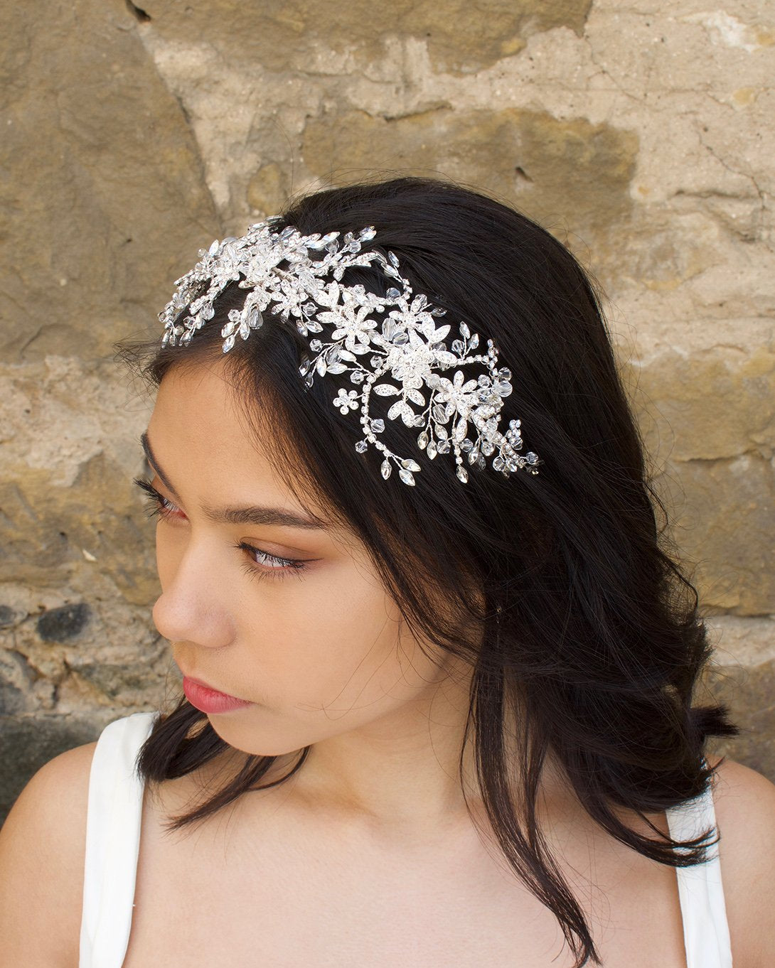 Side view of a bridal model wearing a wide crystal headband for brides with a stone wall backdrop