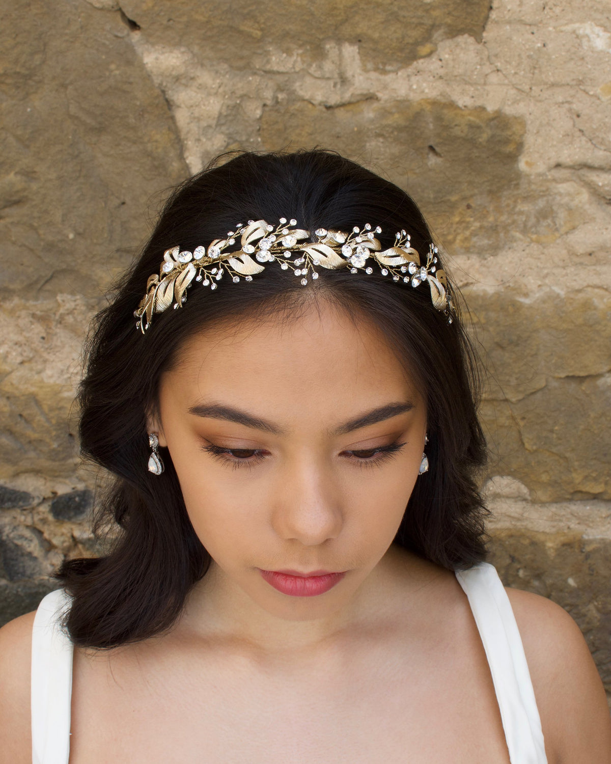 A front view of a model in front of a stone wall wearing a pale gold bridal headband on her forehead.