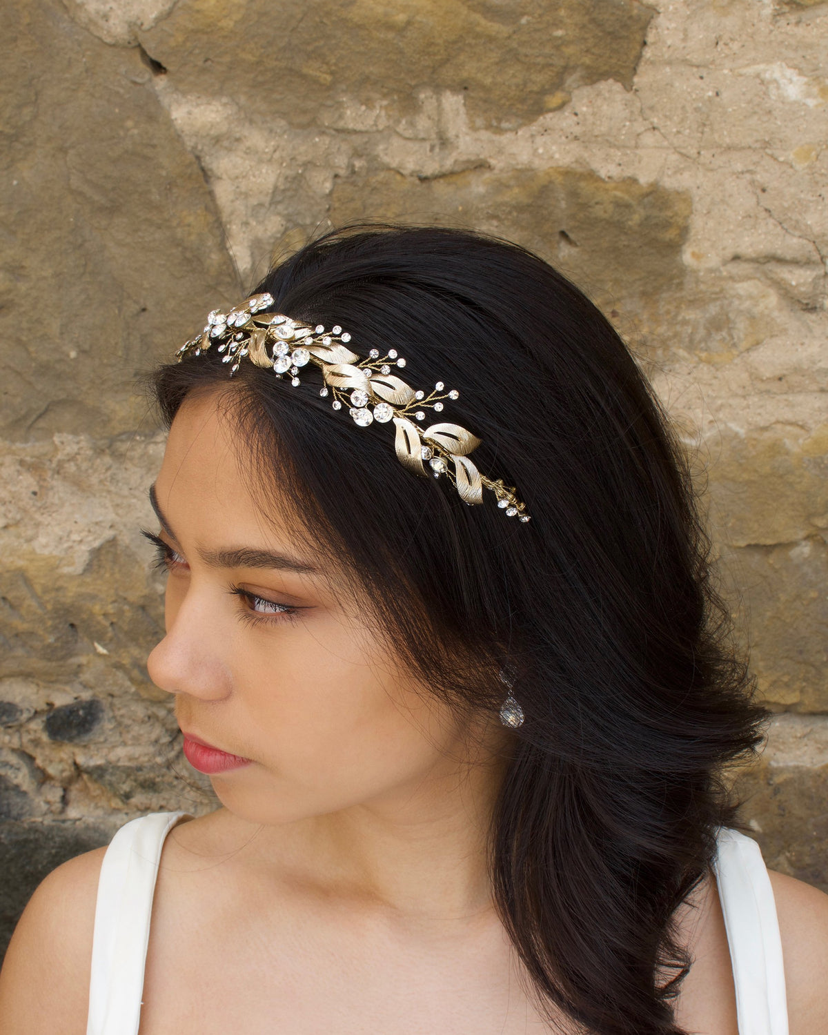 From a side view a Bridal Model with long black hair wears a gold headband of leaf shapes.