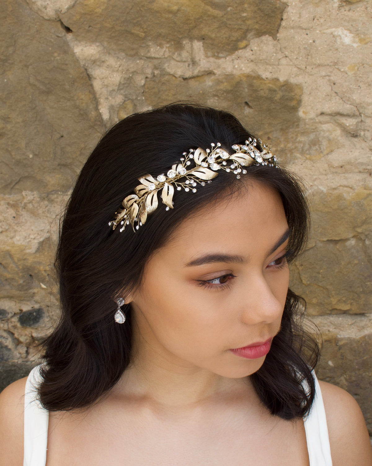 In front of a stone wall a dark hair Bride wears a soft gold bridal headband with tiny crystals