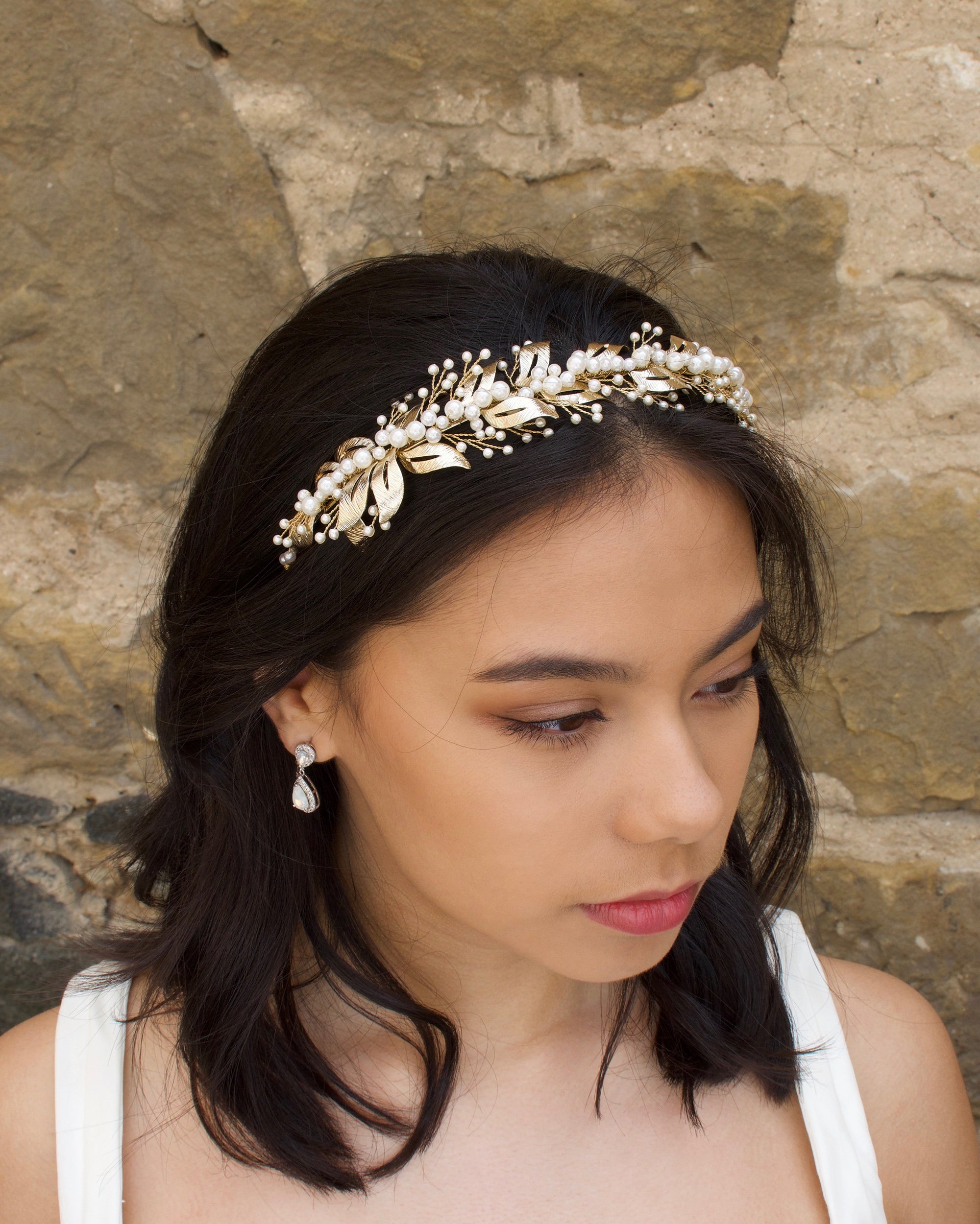 In front of a stone backdrop a dark hair bride wears a headband of pale gold and pearls