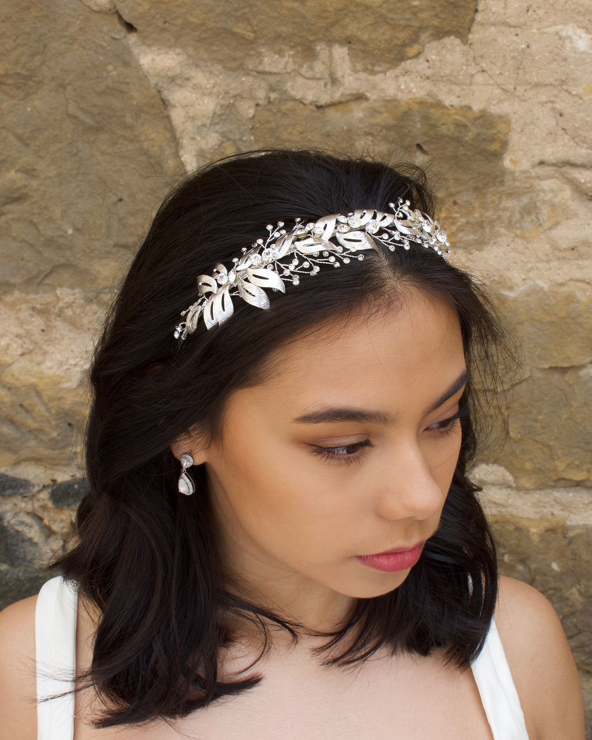 A silver bridal headband is worn by a black haired model with a stone wall in the background