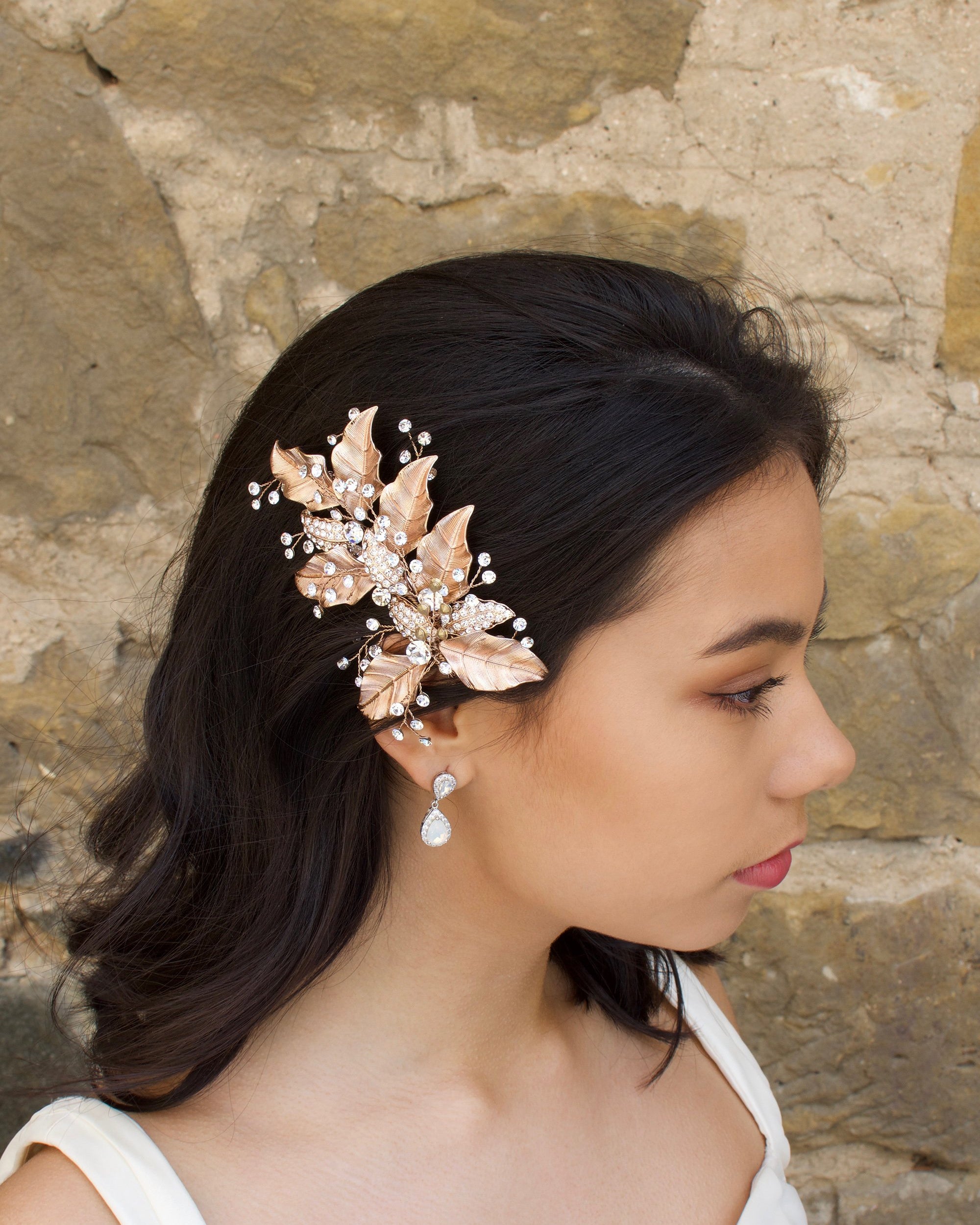 Rose Gold Leaves Side Comb with bright crystal sprigs worn by a dark hair model with a stone wall backdrop.