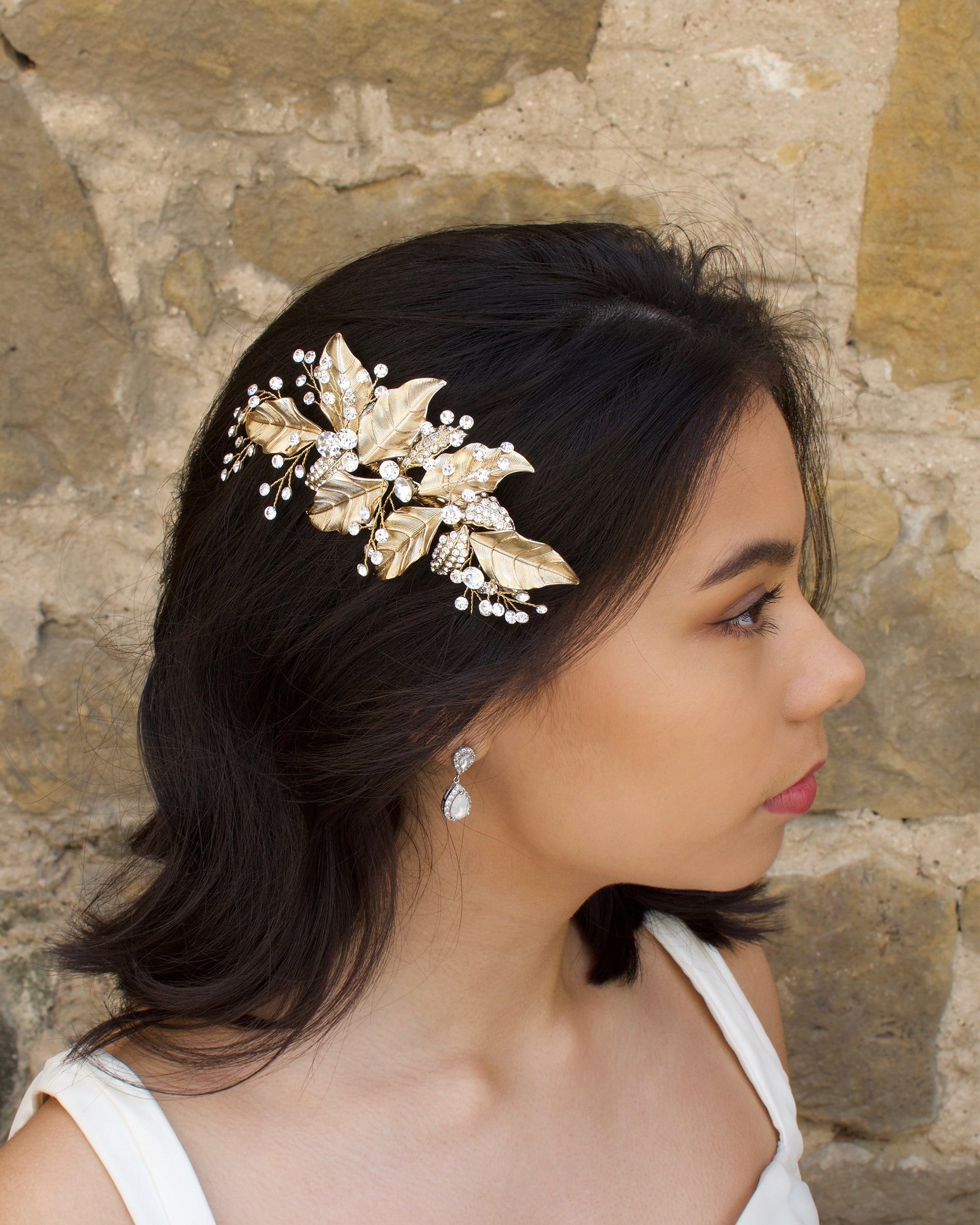 Gold leaf side comb worn by a dark hair bride standing beside a stone wall.