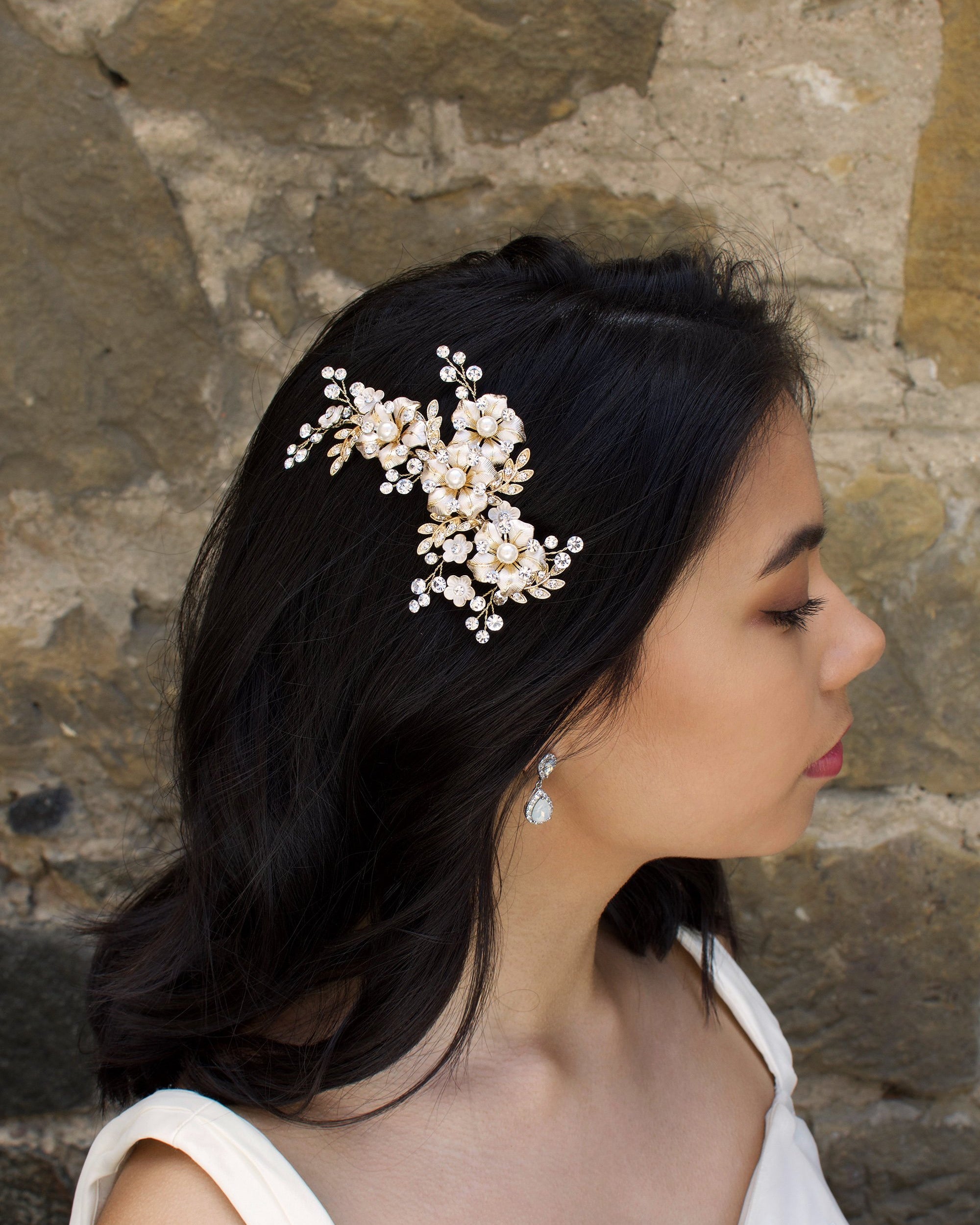 A Dark haired model wears a gold side comb of flowers with a stone wall background