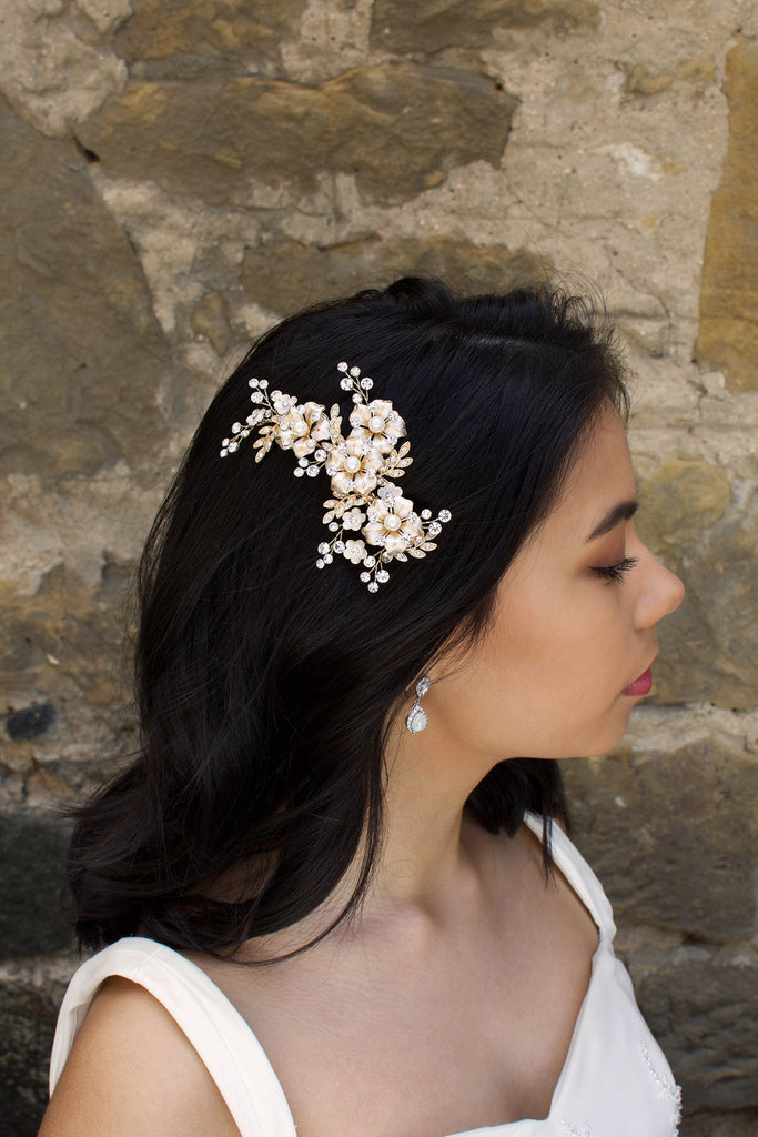 A Dark haired model wears a gold side comb of flowers with a stone wall background