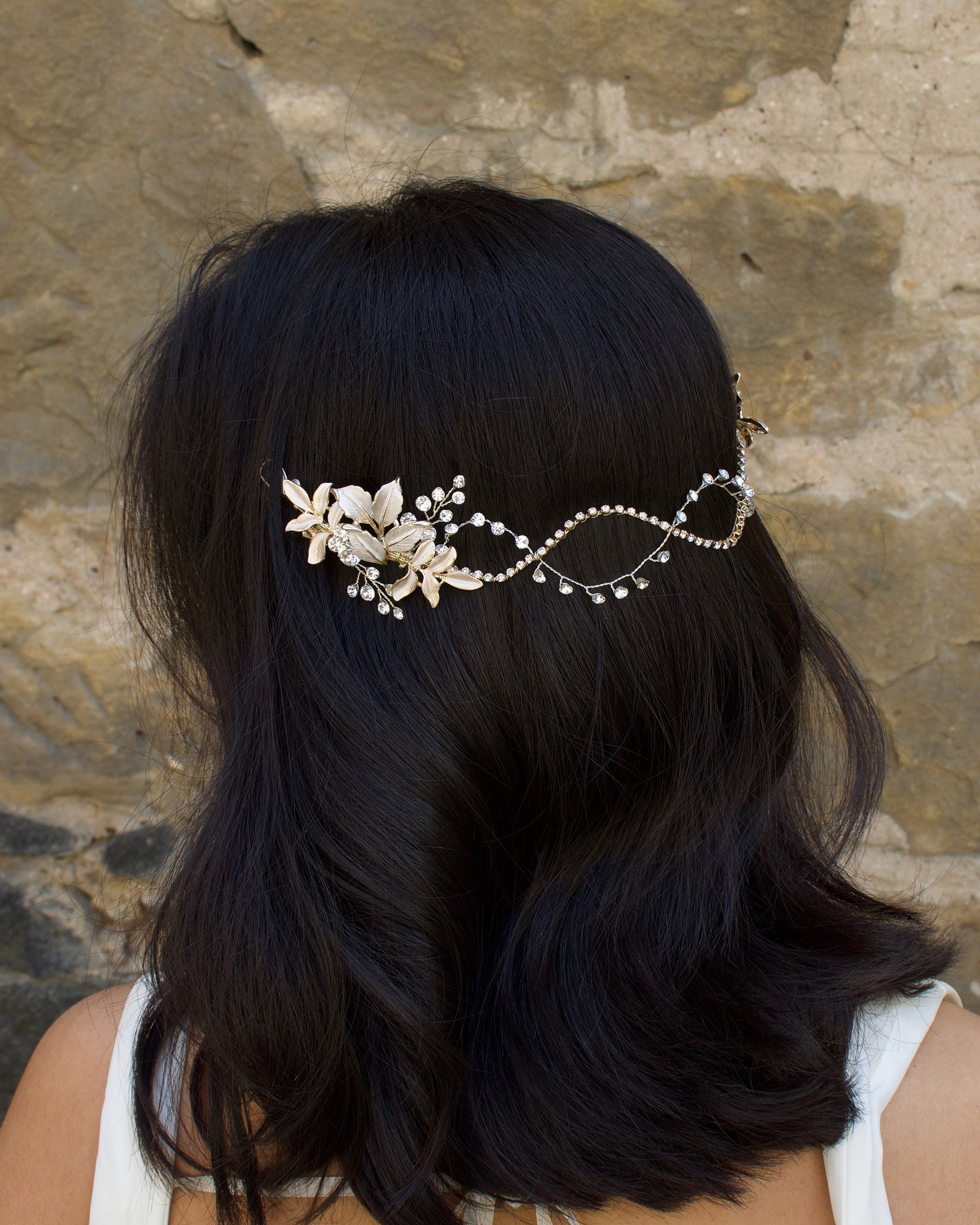 A Black haired model with her hair down wears a Silver soft bridal vine at the back of her head. Stone wall background