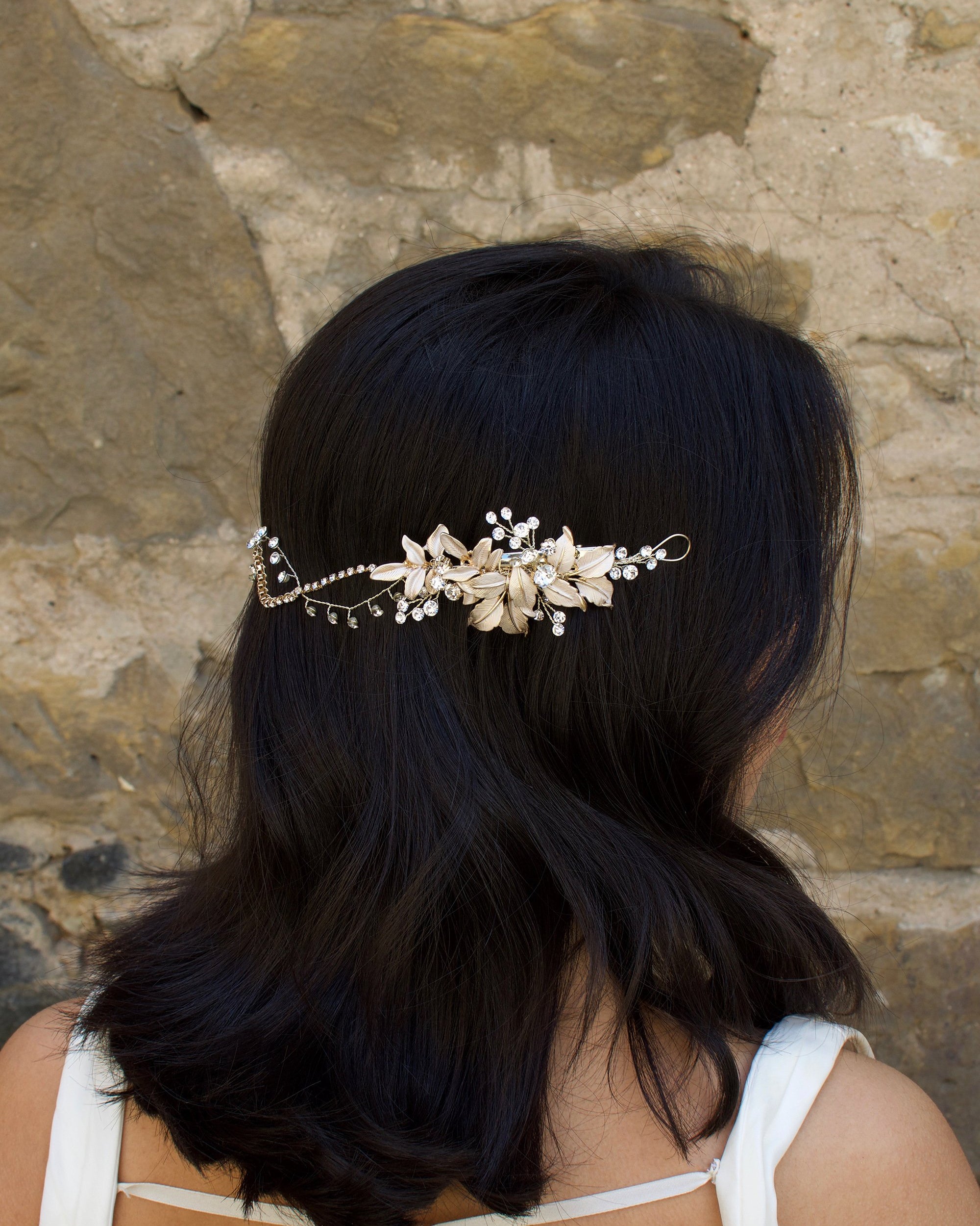 A Black haired model with her hair down wears a soft gold bridal vine at the back of her head. Stone wall background