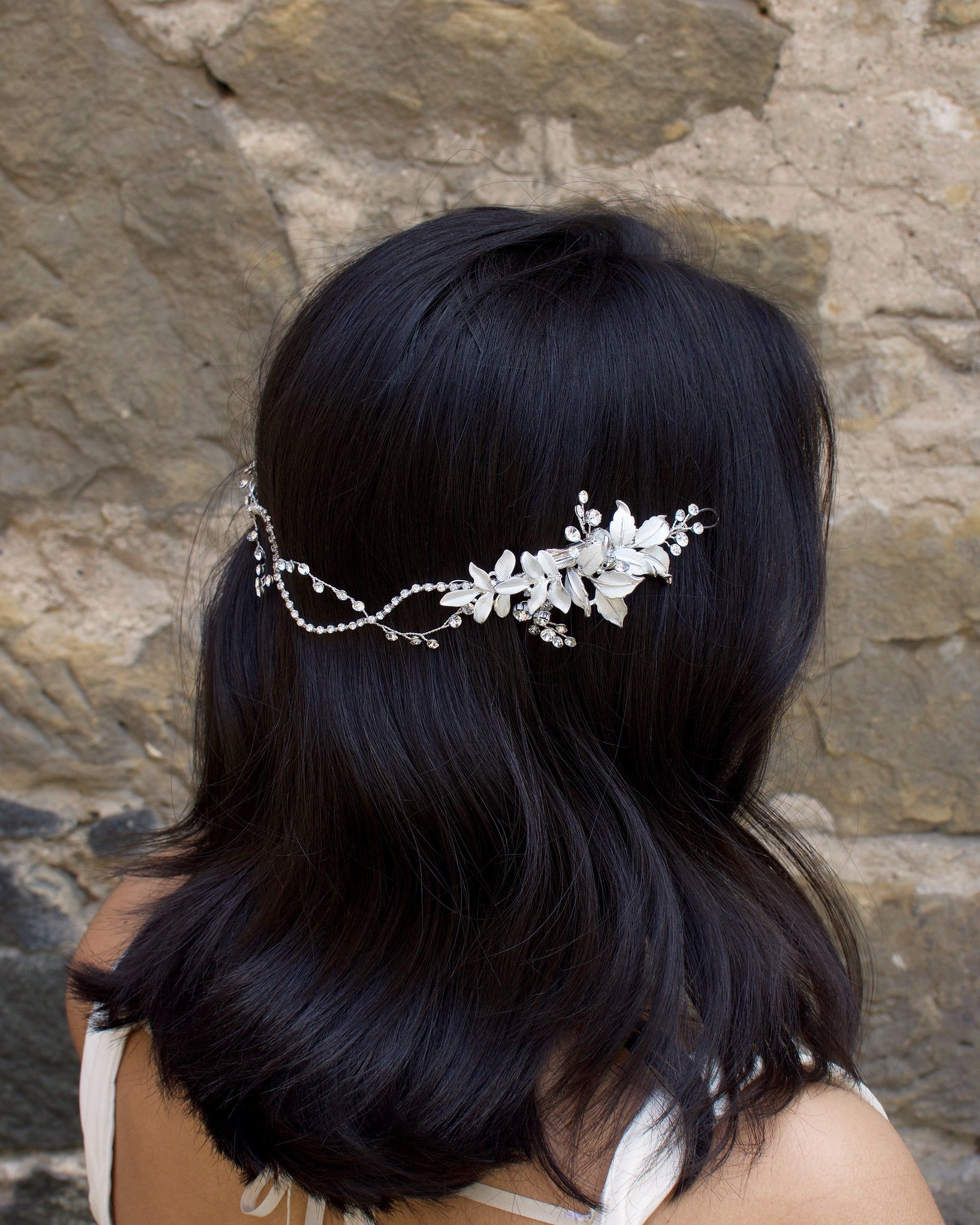 A Black haired model with her hair down wears a soft bridal vine at the back of her head. Stone wall background