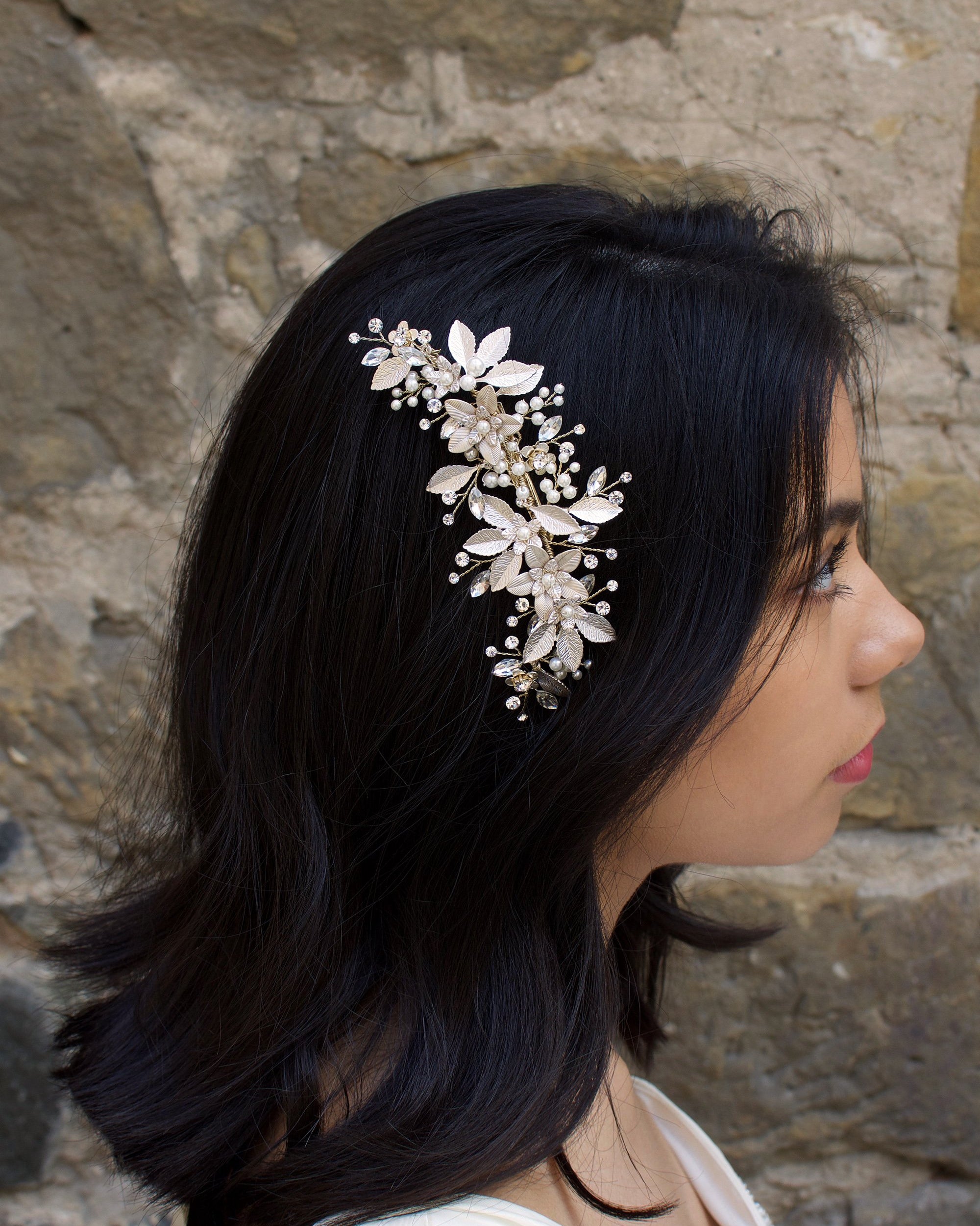 Dark haired model with long hair wears a side comb of pale gold leaves at the side of her head. With a stone wall background