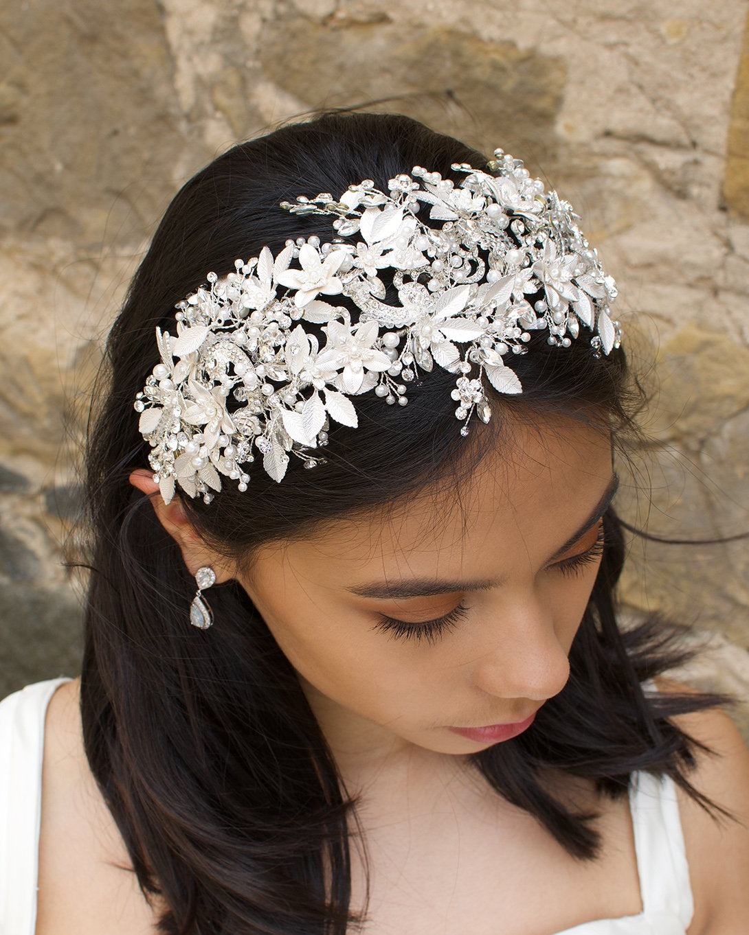 Black hair model looks down wearing a wide bridal headpiece with many leaves. Stone wall is behind