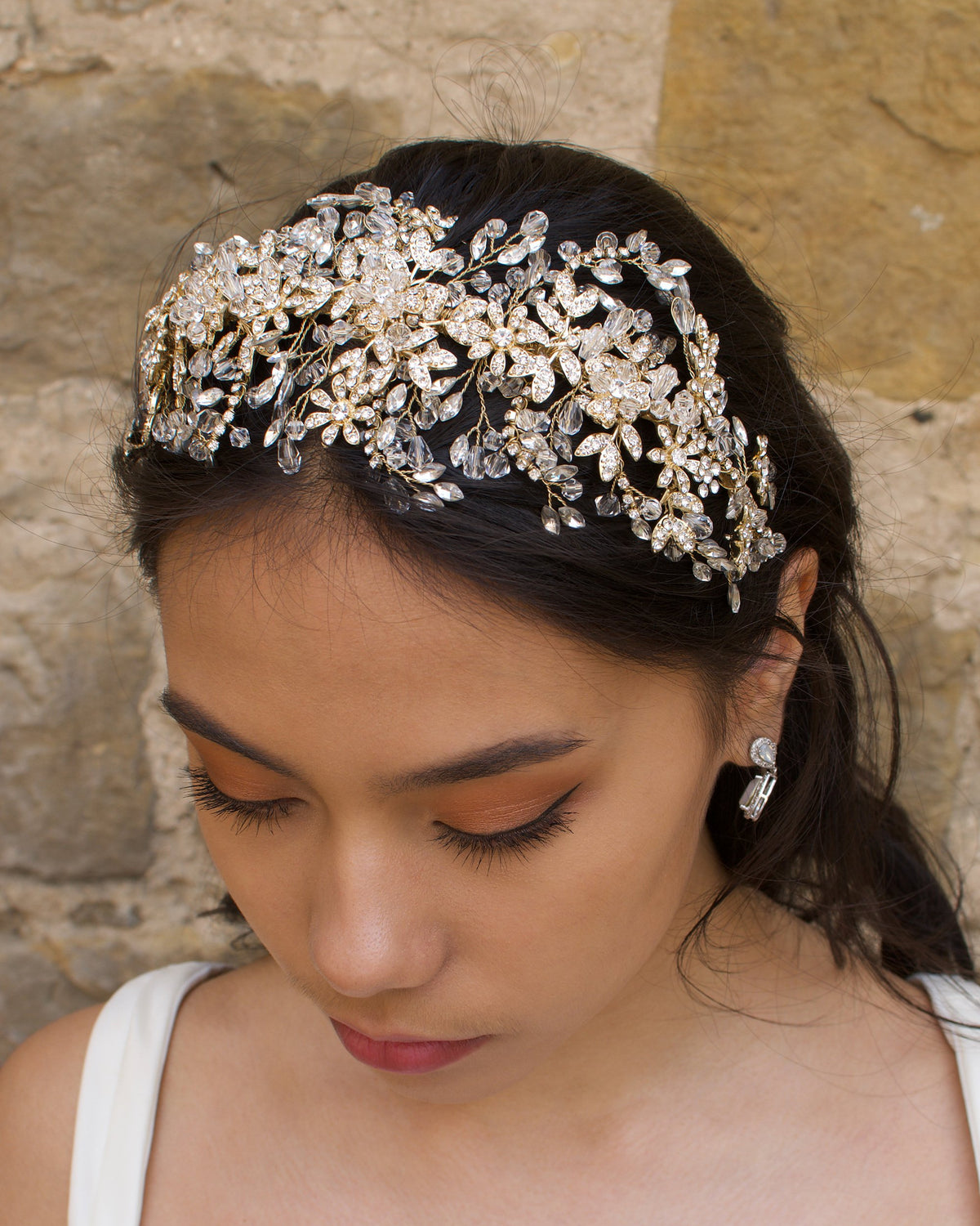 A model wears a wide golden headband across the front of her head with a stone wall background