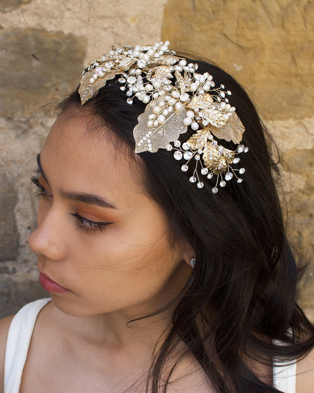 A wide gold headband worn by a dark hair bride with a stone wall background