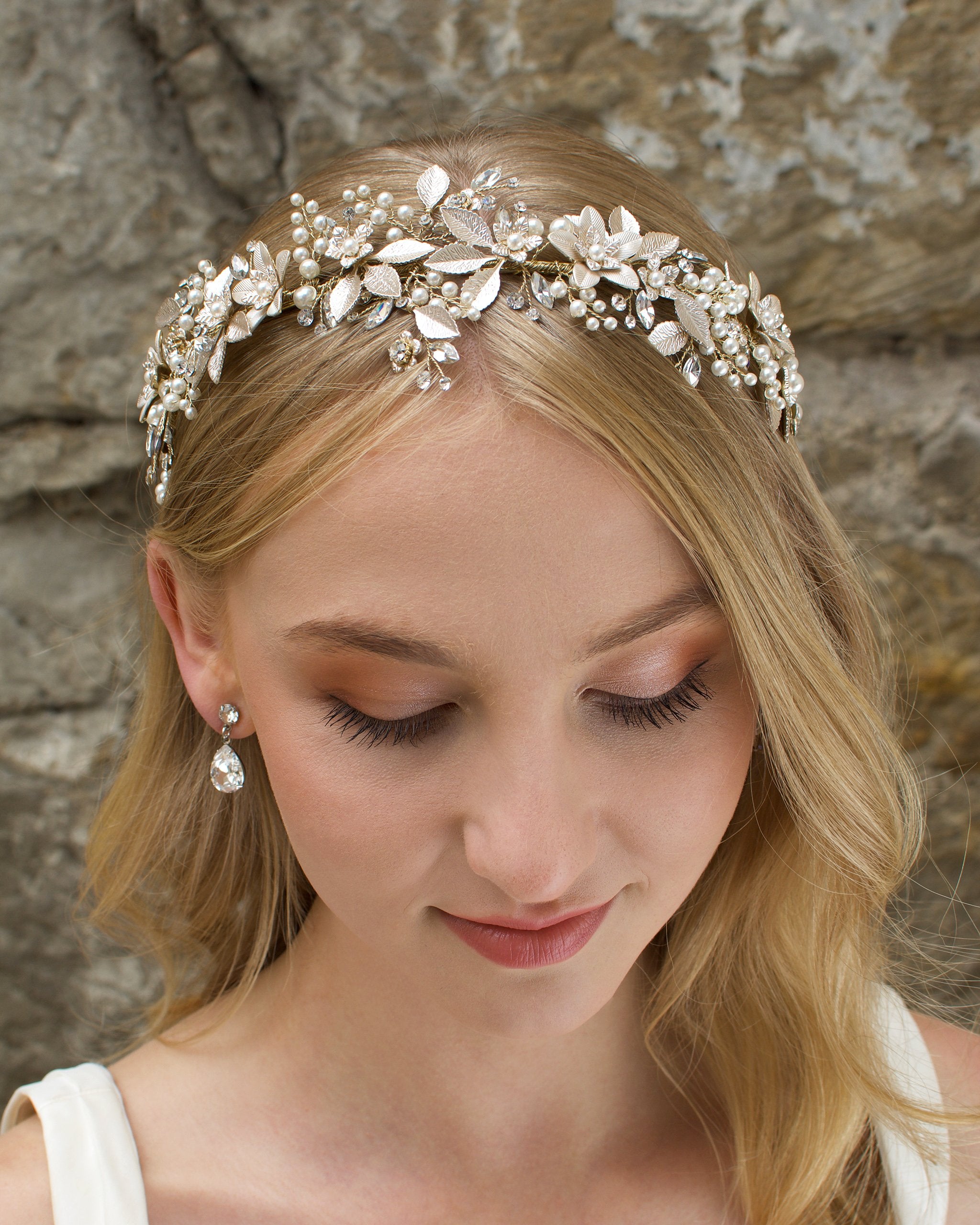 A model Bride with blonde hair wears a gold bridal crown at the front of her head with a stone wall behind