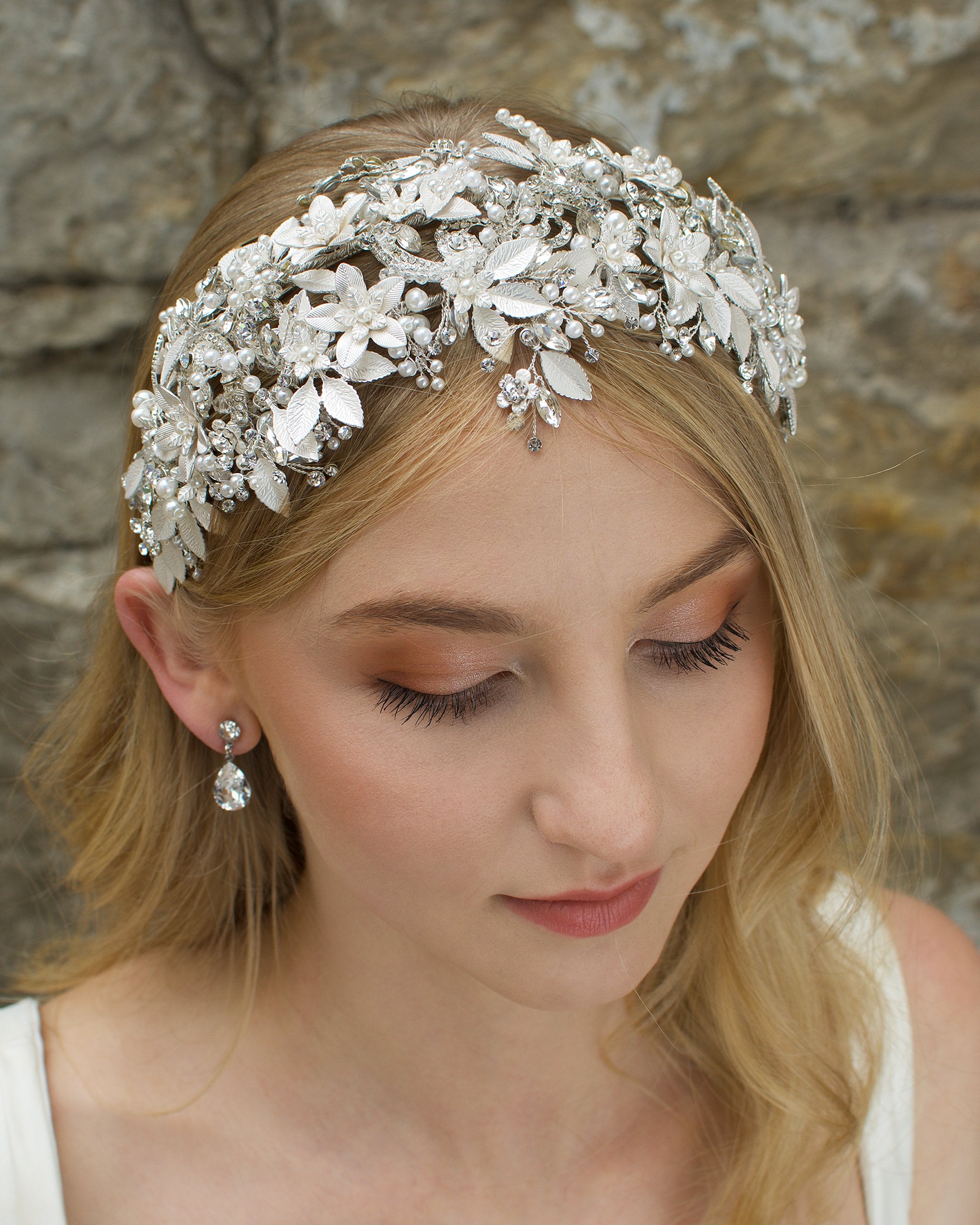 Model facing the camera wears a wide silver leaves bridal headband and the background is an old stone wall.