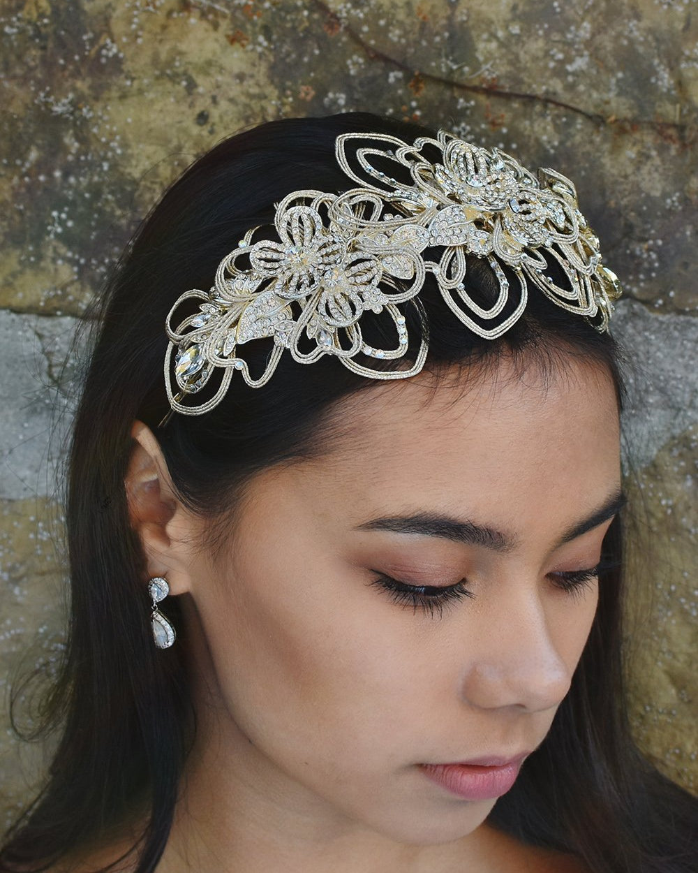 Dark haired model wearing a matt gold bridal headband at the front of her head. There is a stone wall behind her.