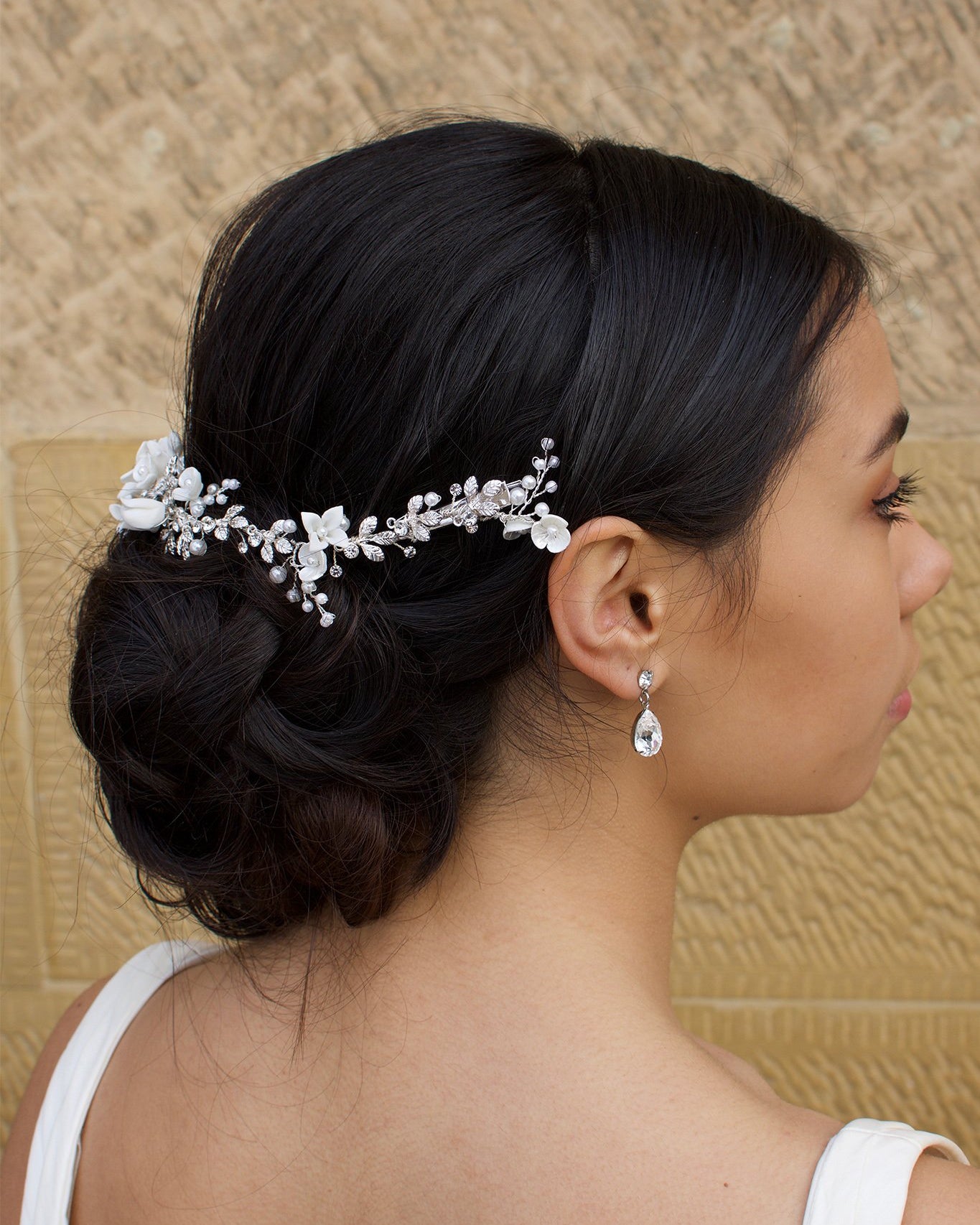 Side view of a black haired Bride wears a porcelain flowers silver vine around the back of her head with a stone wall backdrop