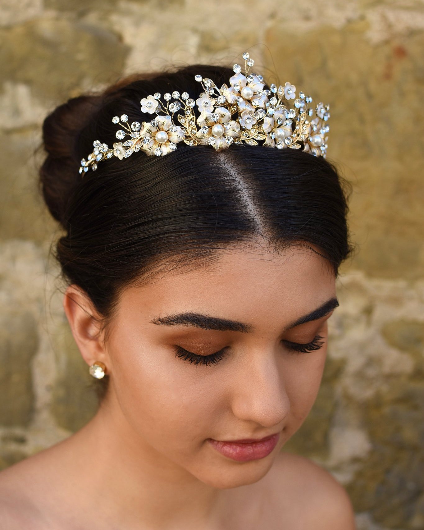 A dark model wears a gold tiara with pearl flowers against a stone wall background.
