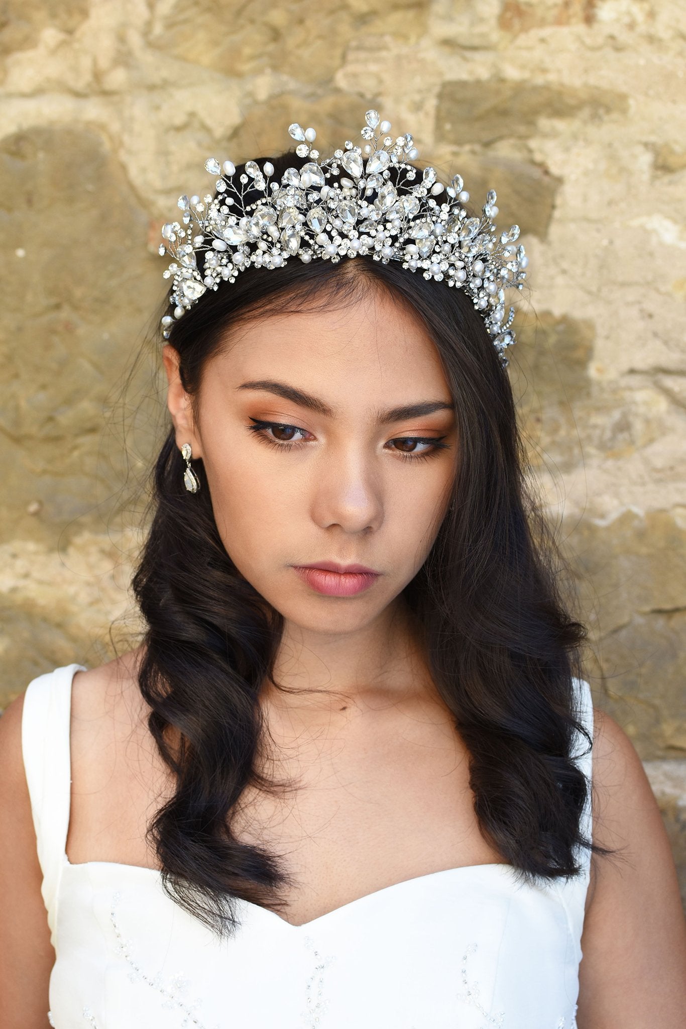 A model Bride wears a high Tiara of real pearls and crystals high on her head. She has dark hair and behind her is a stone wall.