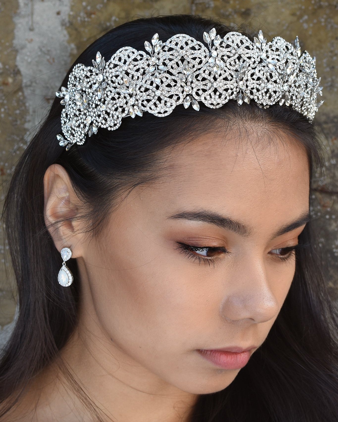 A wide crystal crown is worn by a dark haired model standing in front of a stone wall