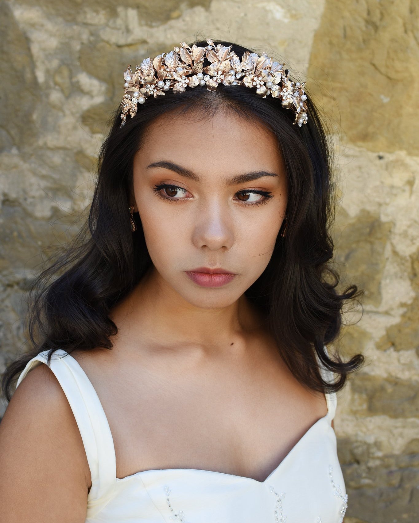 Bridal Model wears a pale rose gold tiara in her dark hair standing against an old stone wall